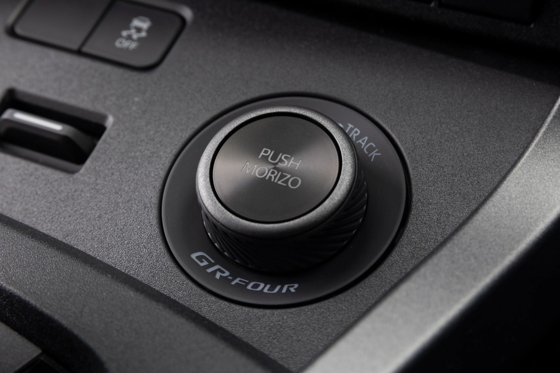 Black textured car control knob labeled "PUSH MORIZO" with "GR-FOUR" and "TRACK" markings on a matte black panel.