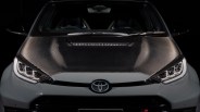 Front view of a white Toyota car with a black carbon fiber hood and illuminated headlights.