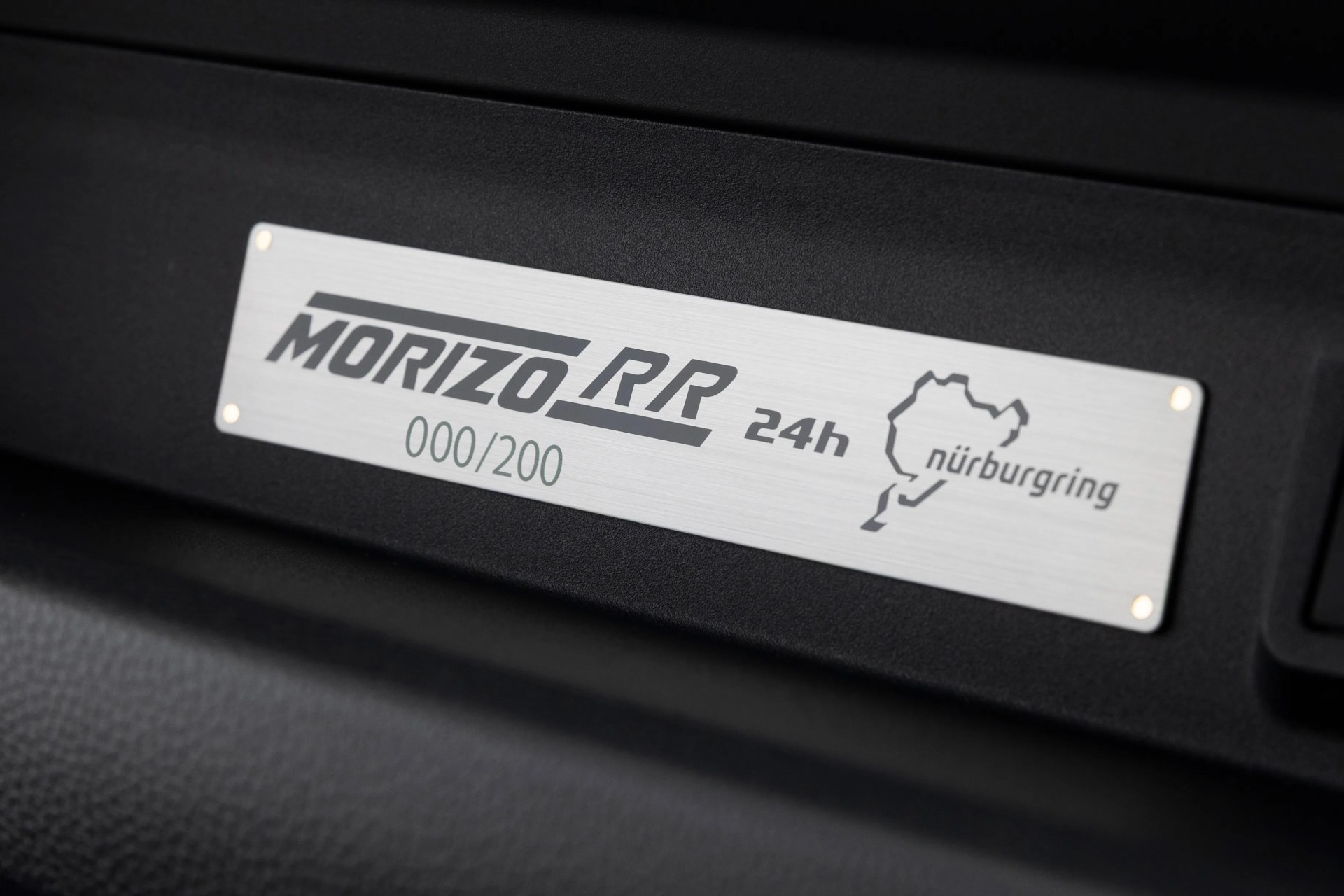 Silver metal plaque with "MORIZO RR 24h Nürburgring 000/200" mounted on a black textured surface.