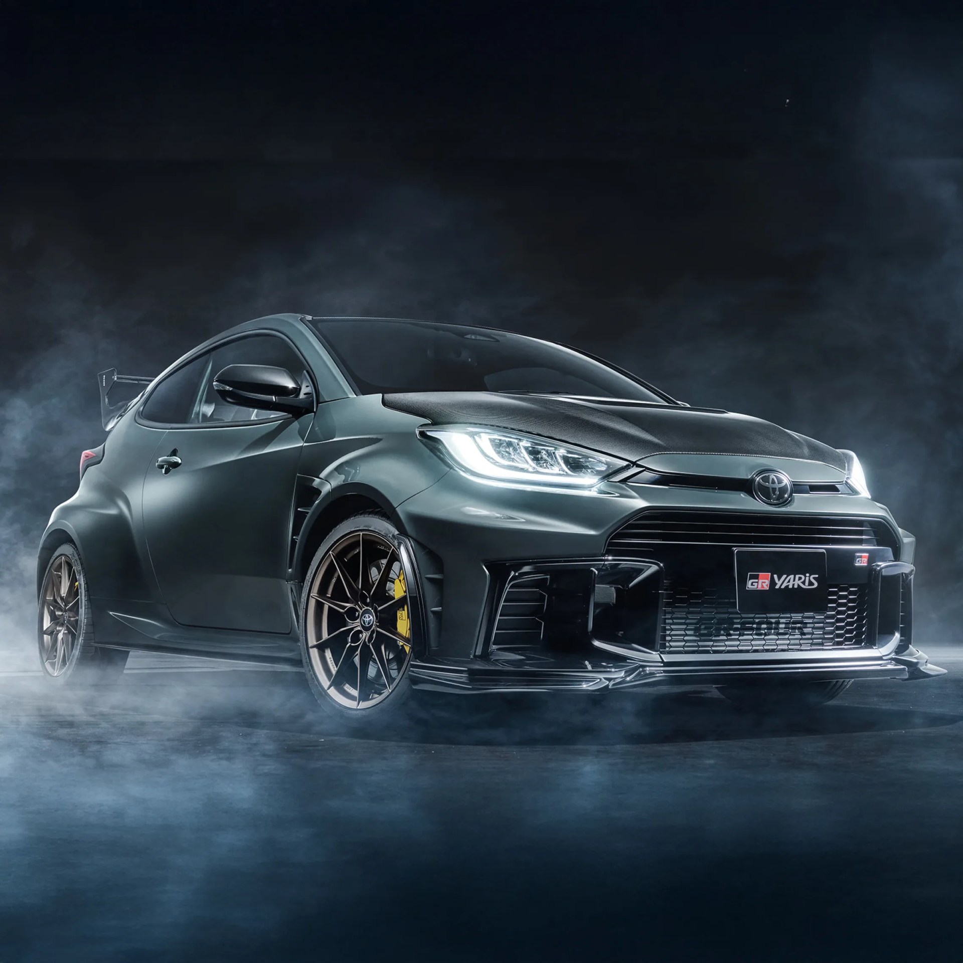 Dark gray Toyota GR Yaris with illuminated headlights and bronze wheels in smoky setting.