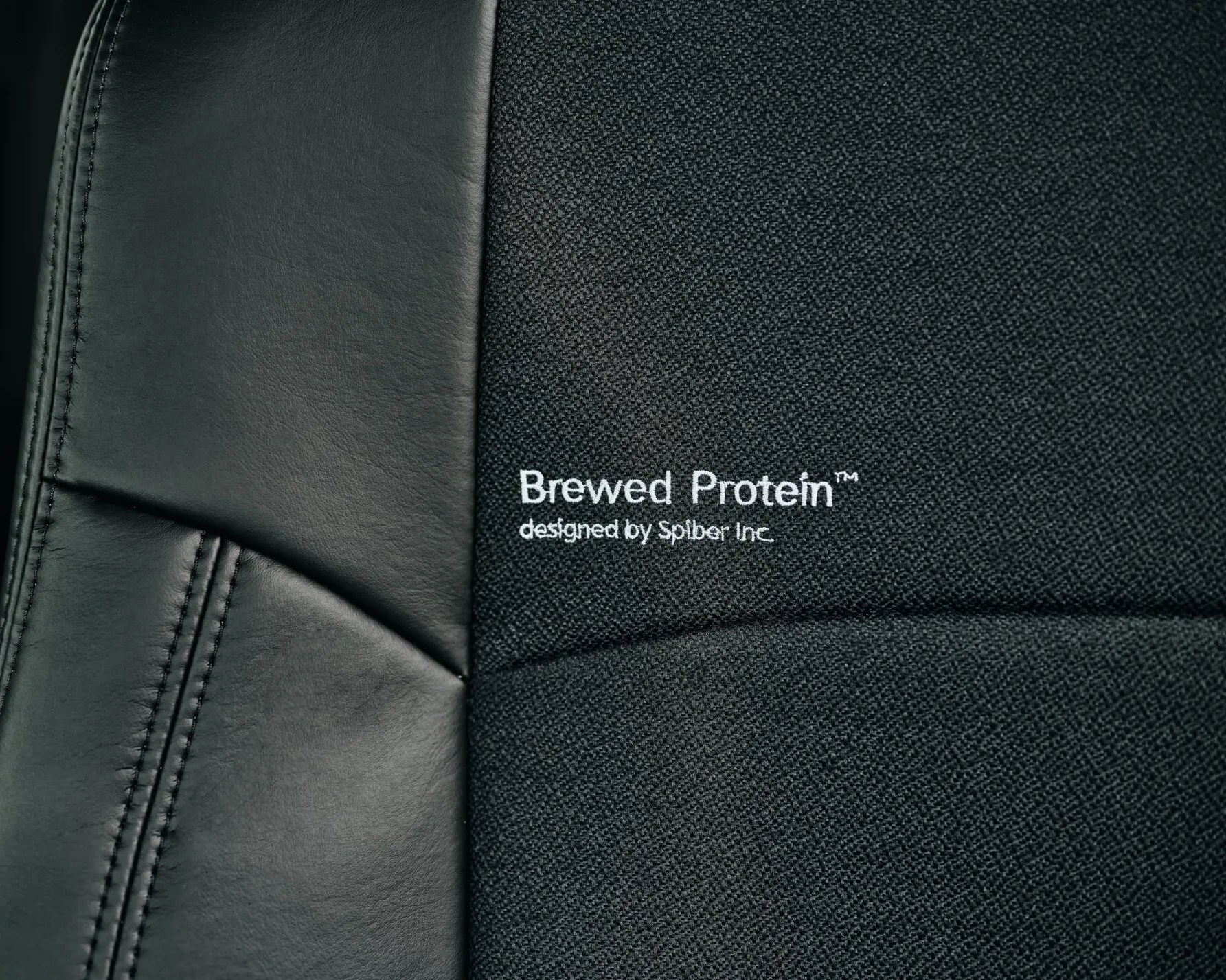 Close-up of black leather and fabric chair upholstery with white embroidered text "Brewed Protein&trade; designed by Spiber Inc.
