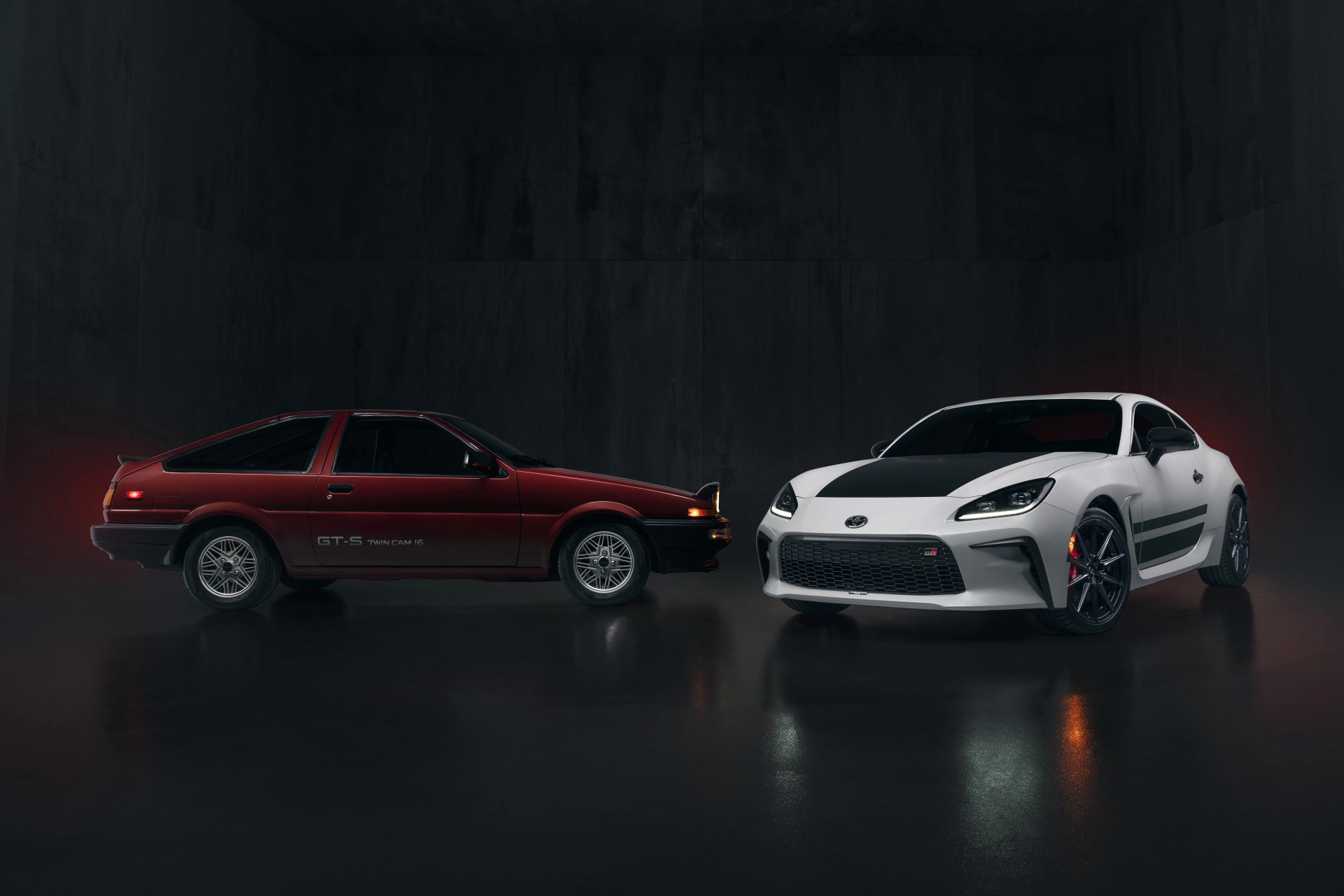 Side view of a red vintage Toyota GT-S and a front three-quarter view of a white modern Toyota GR 86 in a dark studio.