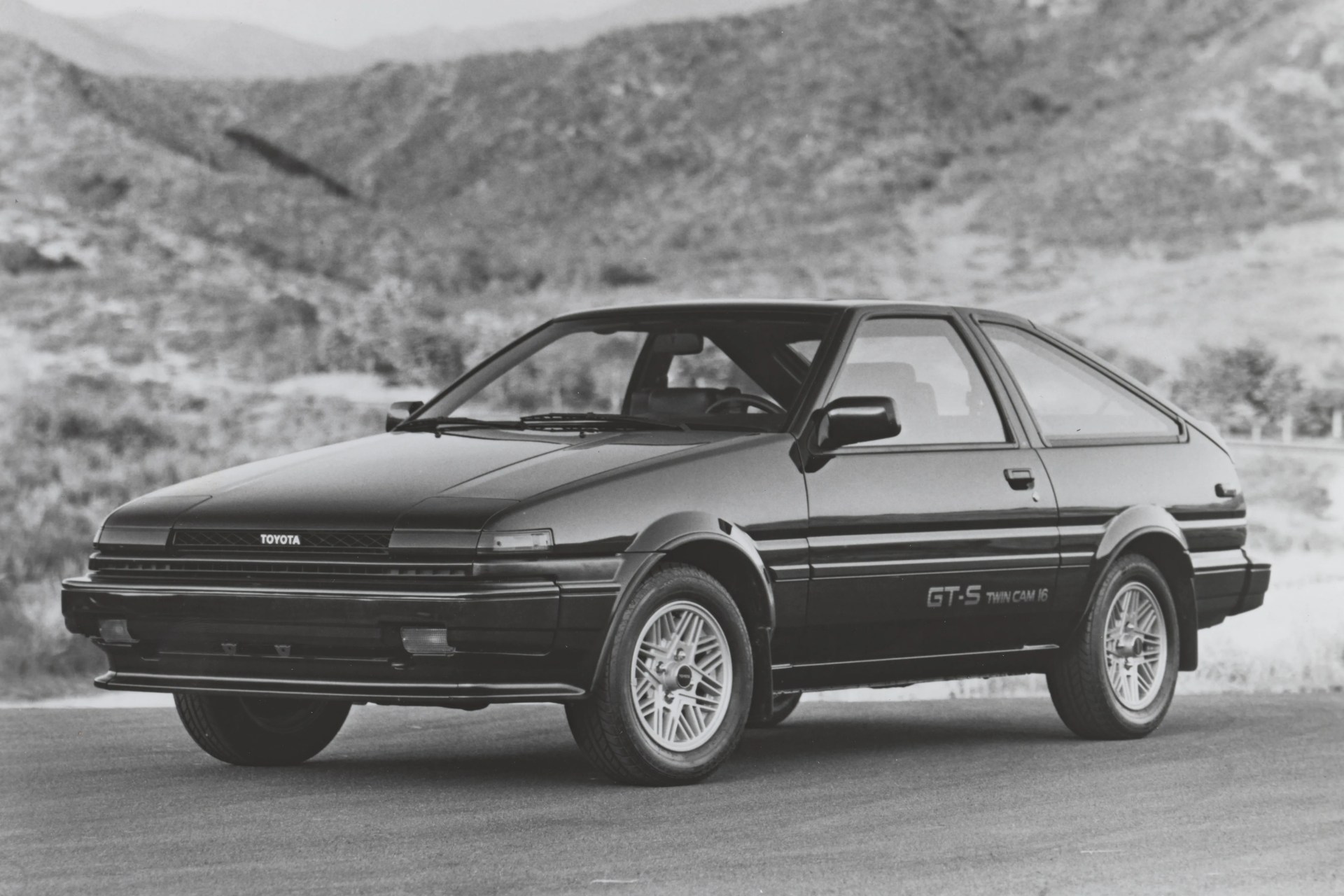 Black Toyota GT-S Twin Cam 16 hatchback parked on a road with mountainous background.