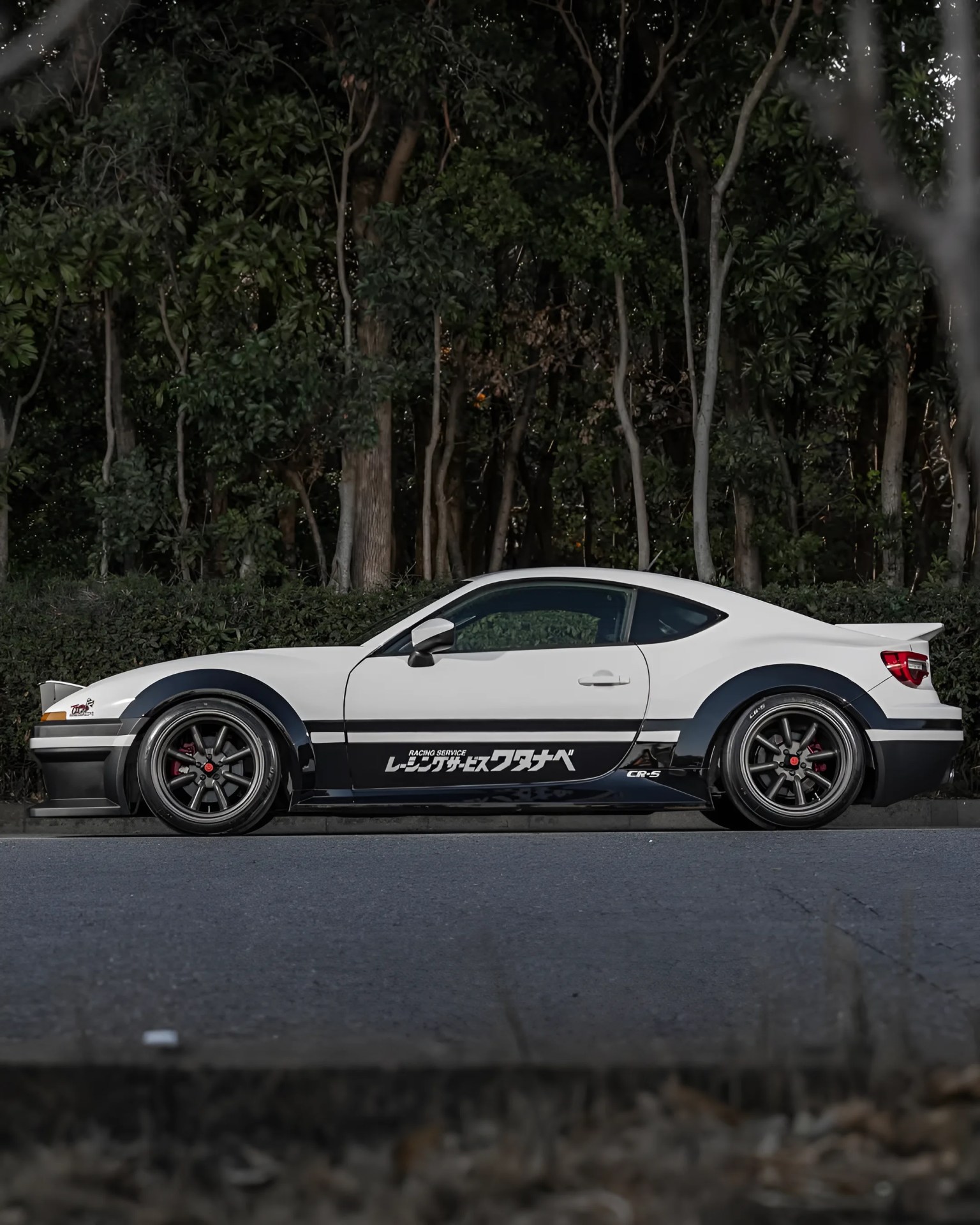 White and black modified sports car with Japanese text decals parked on a road in front of trees.
