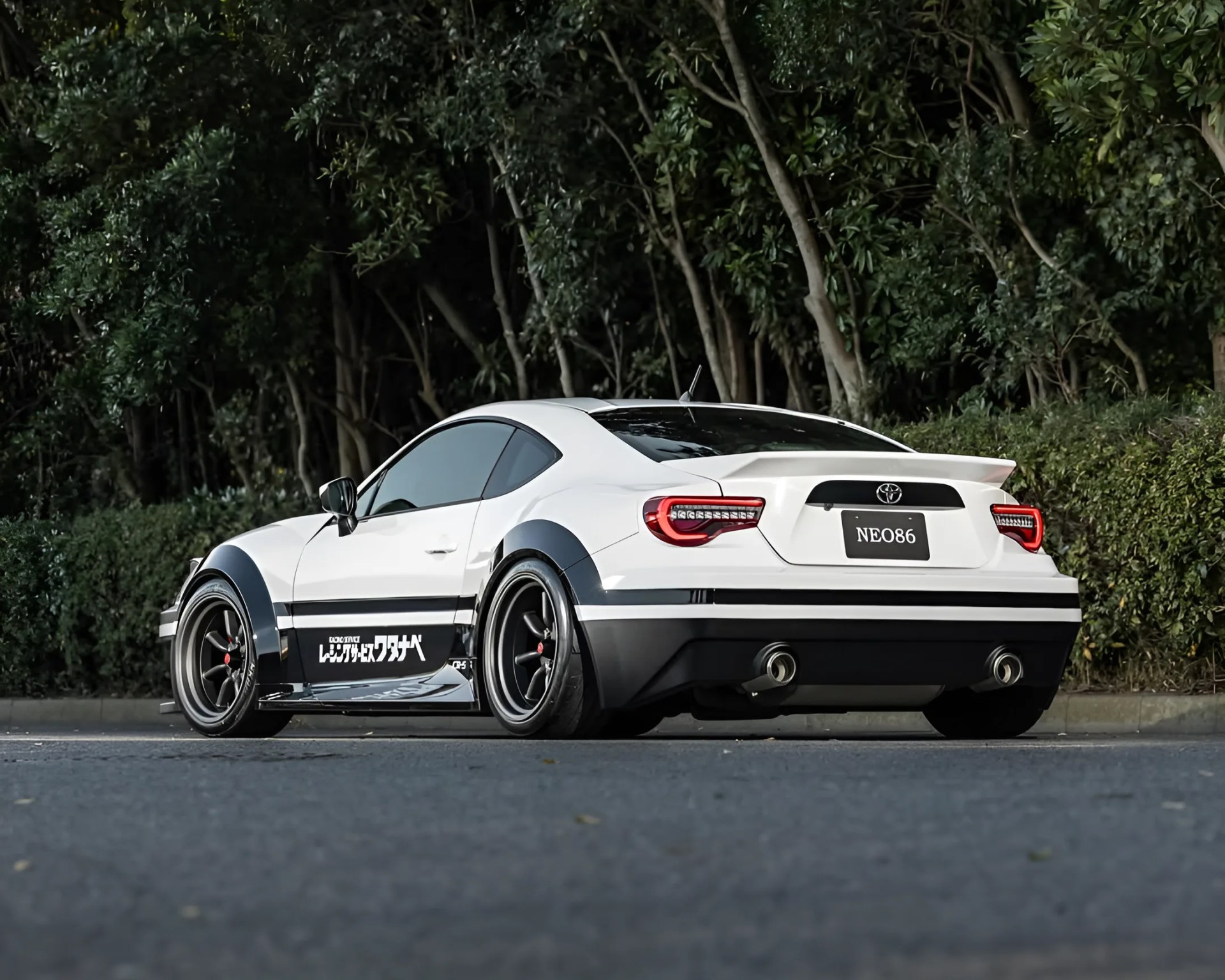 White Toyota sports car with black wide fenders, black wheels, and Japanese decals parked on a road near trees.