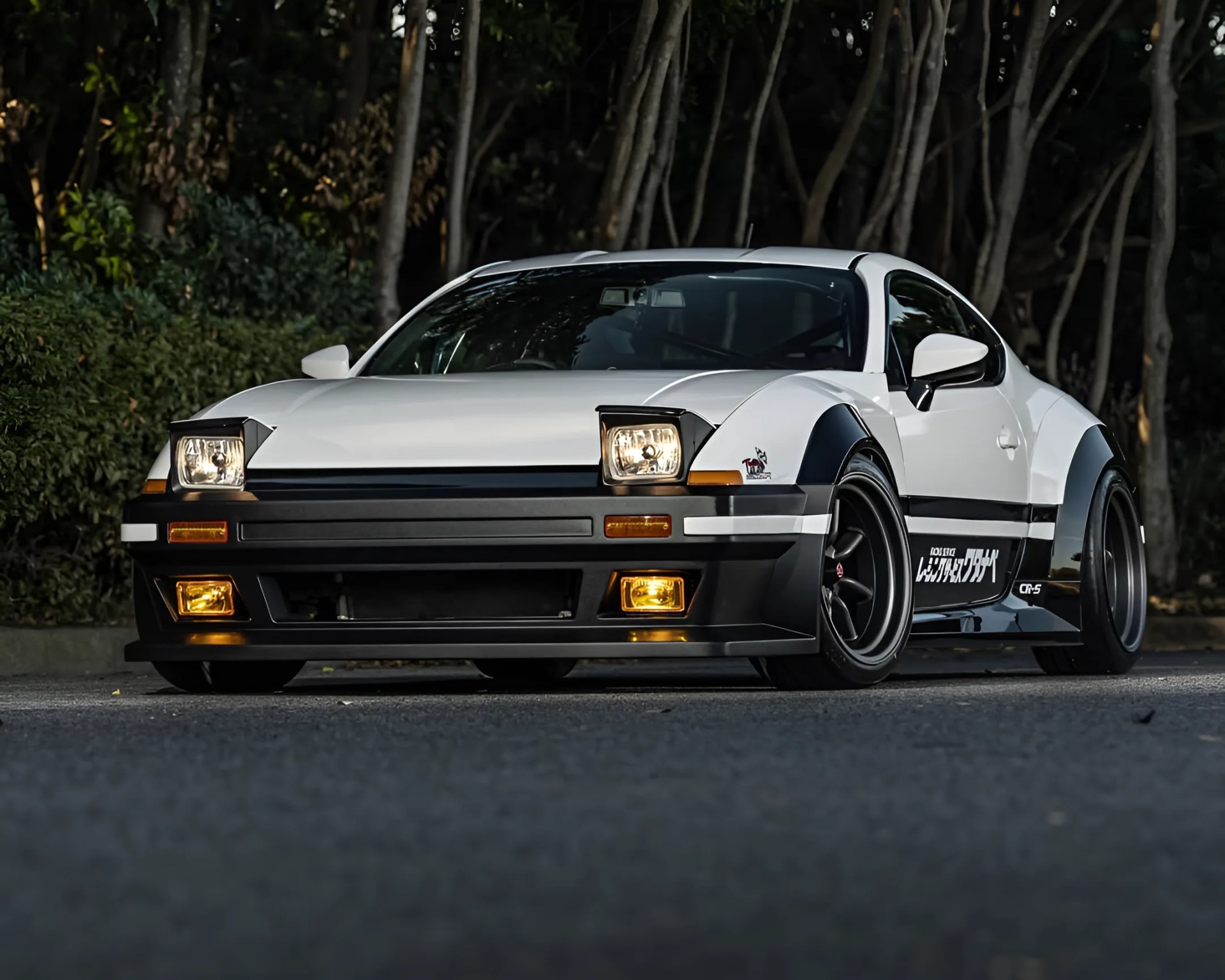 White and black wide-body sports car with pop-up headlights and Japanese decals parked on a road with trees in the background.