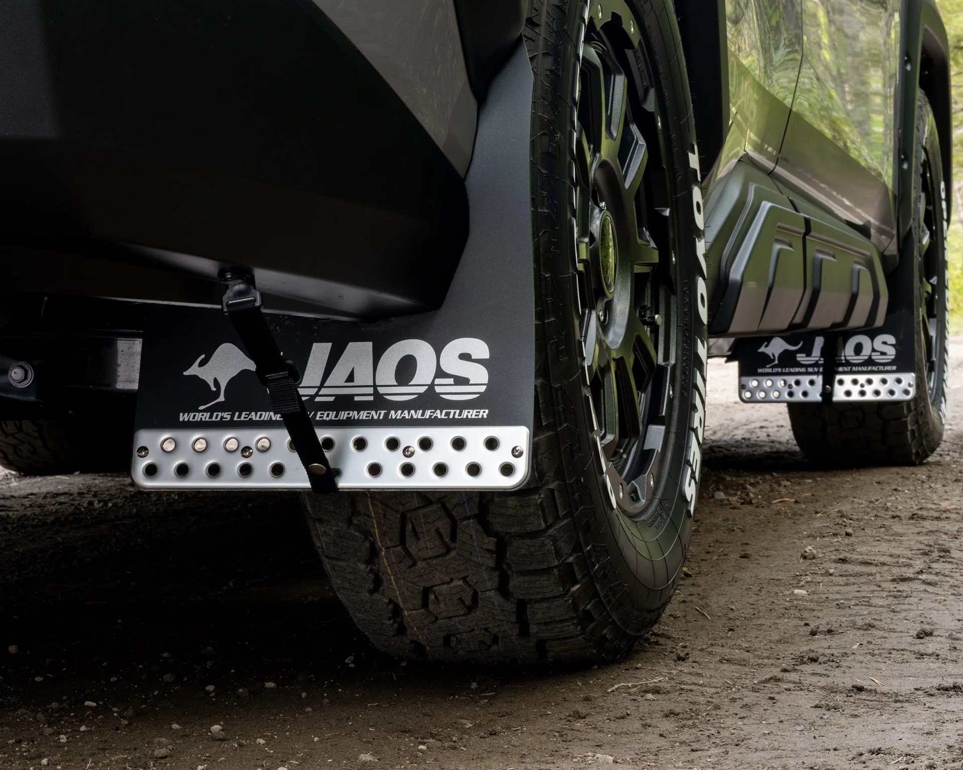 Close-up of black JAOS mud flaps with metal reinforcement plates installed behind rugged off-road tires on a green vehicle.