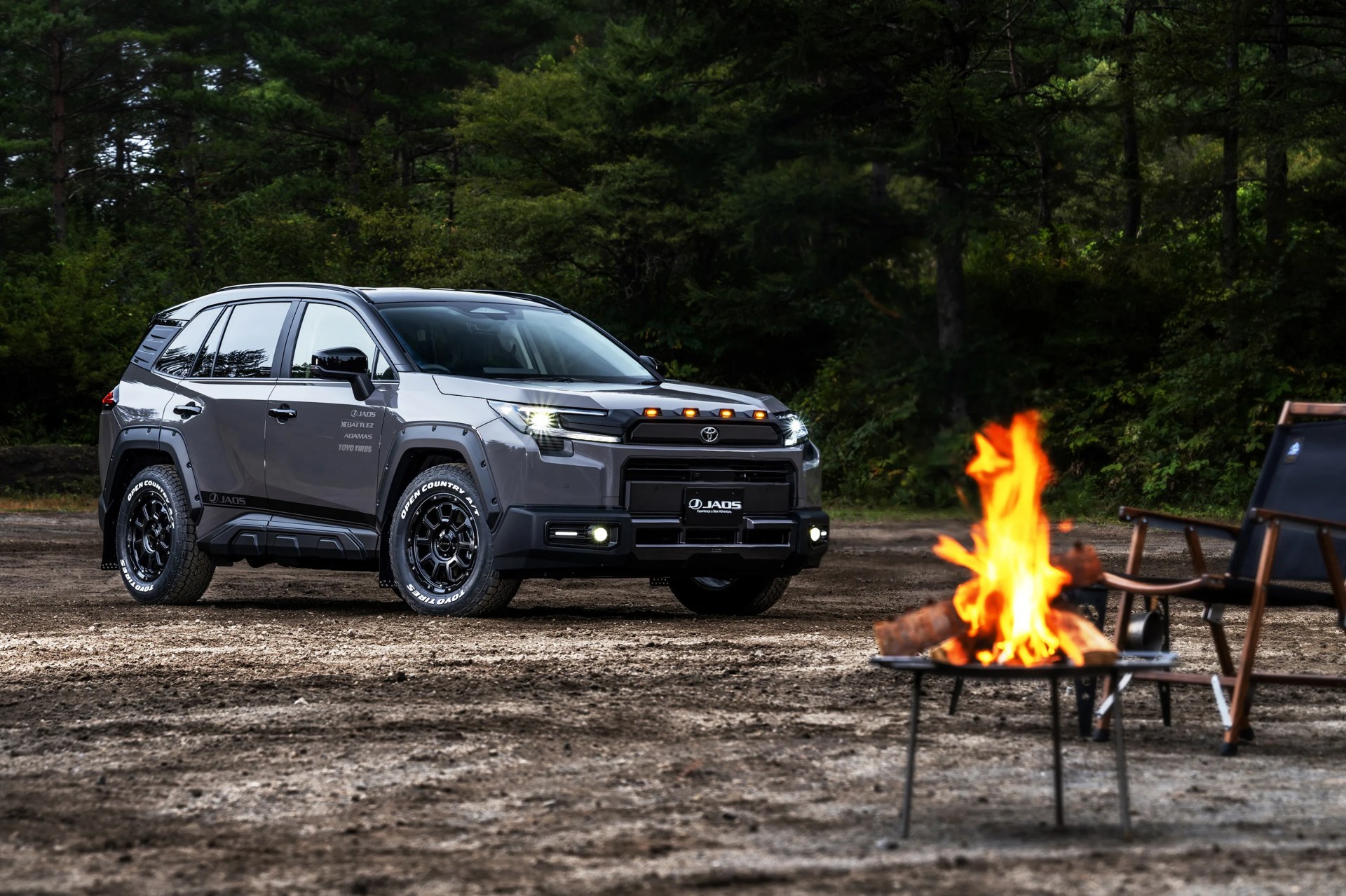 Gray Toyota SUV with off-road tires parked on dirt near a campfire and wooden chairs in a forest setting.
