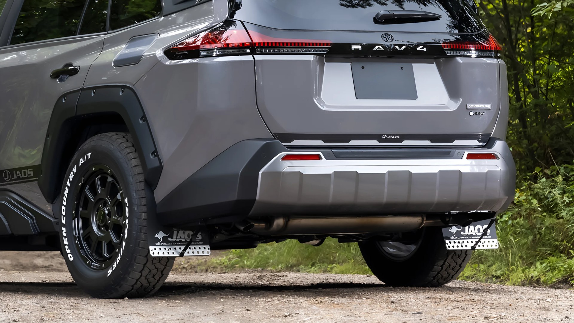 Rear view of a gray Toyota RAV4 Adventure with black off-road tires and JAOS mud flaps on a dirt road.