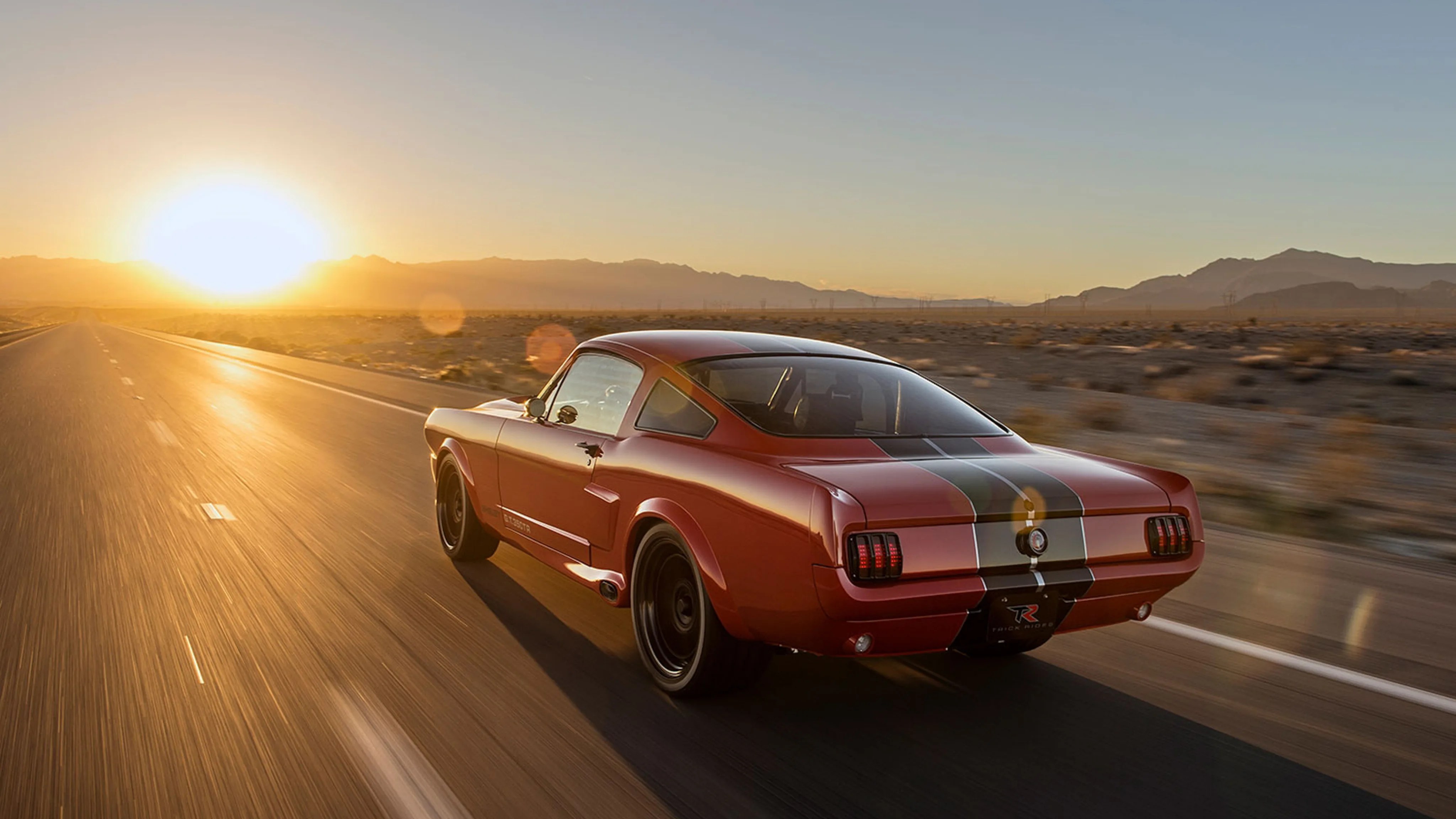 Red vintage muscle car with black racing stripes driving on a desert highway at sunset.
