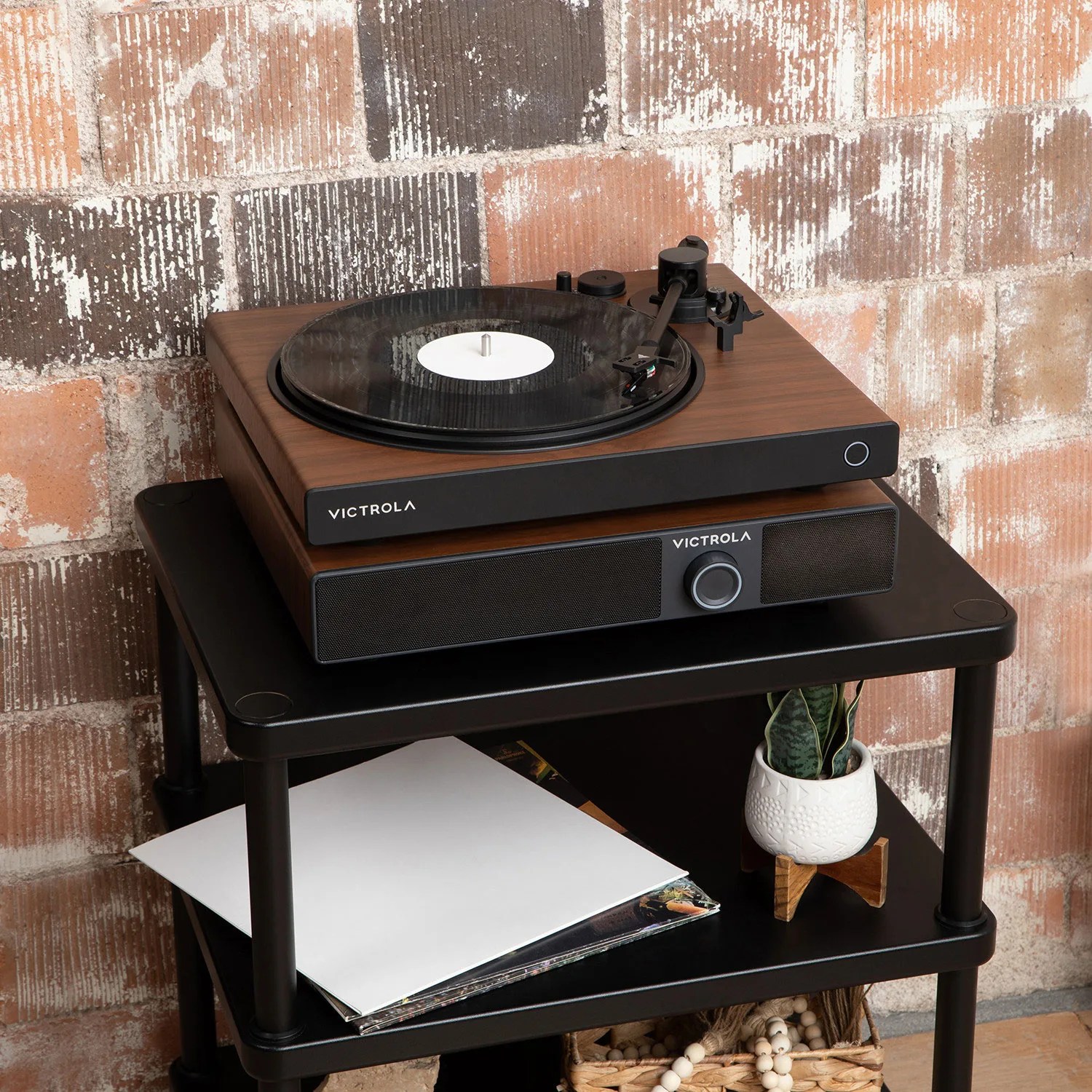 Victrola wooden turntable with built-in speaker on a black two-tier cart against a brick wall.