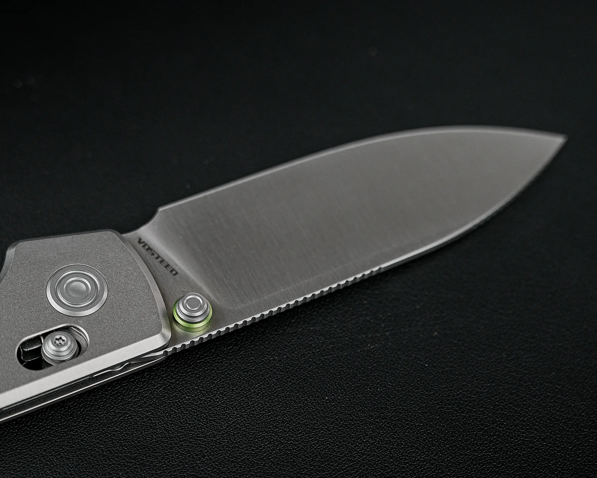 Close-up of a partially serrated stainless steel folding knife blade with a thumb stud and textured handle on a black surface.