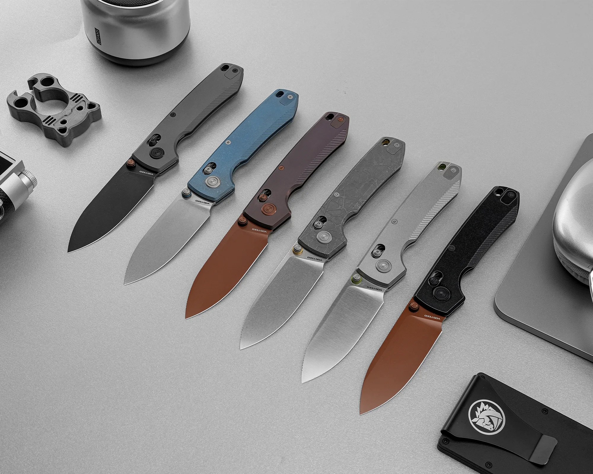 Six folding knives with various handle colors and blade finishes arranged in a row on a gray surface.