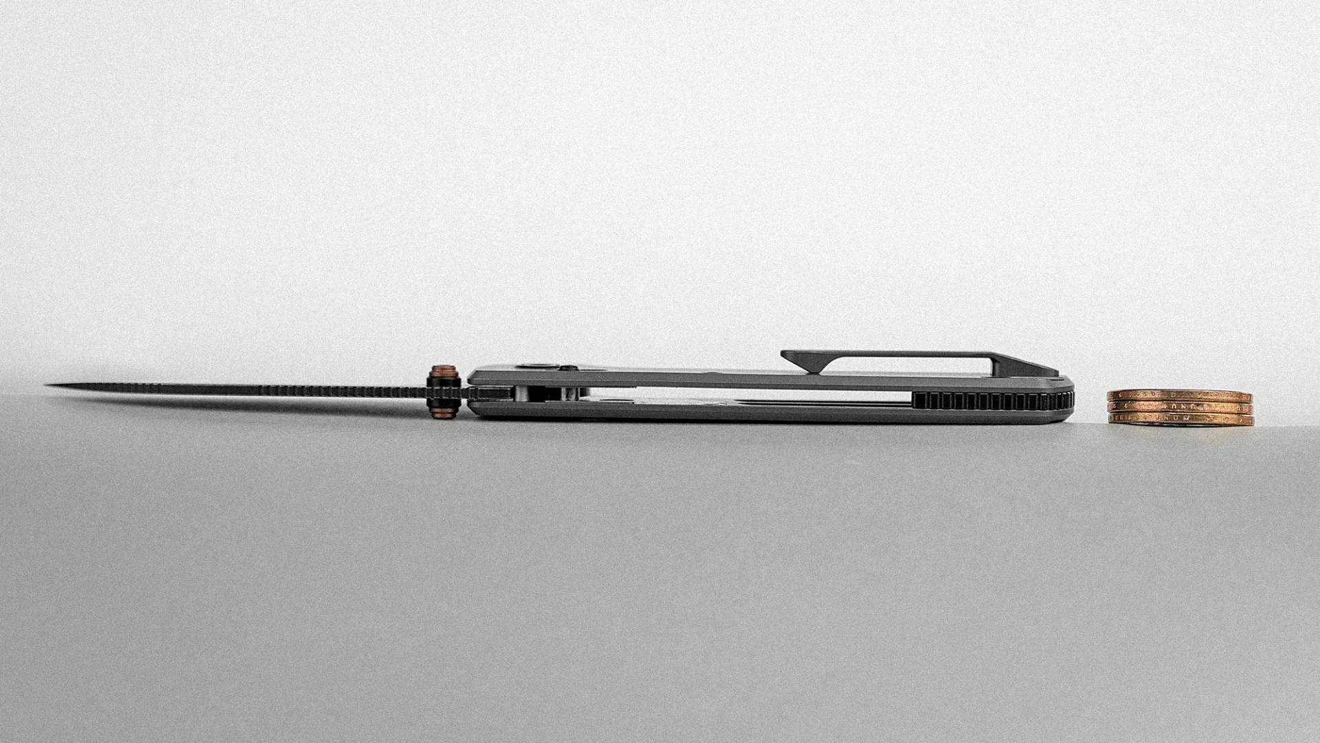 Folded black pocket knife with a clip lying flat next to a small stack of copper coins on a gray surface.