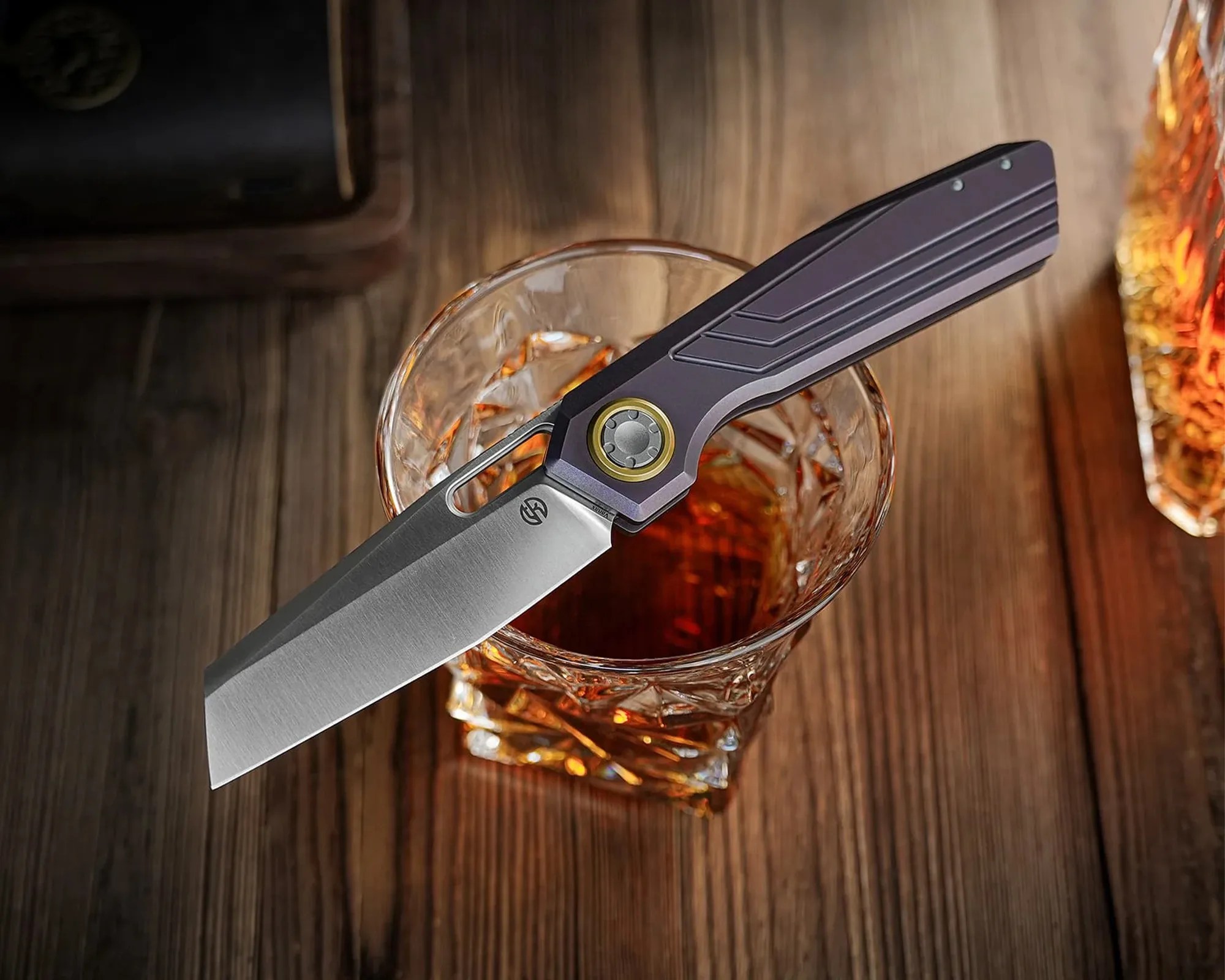 Folding knife with a gray blade and purple handle resting on a glass of amber liquid on a wooden surface.