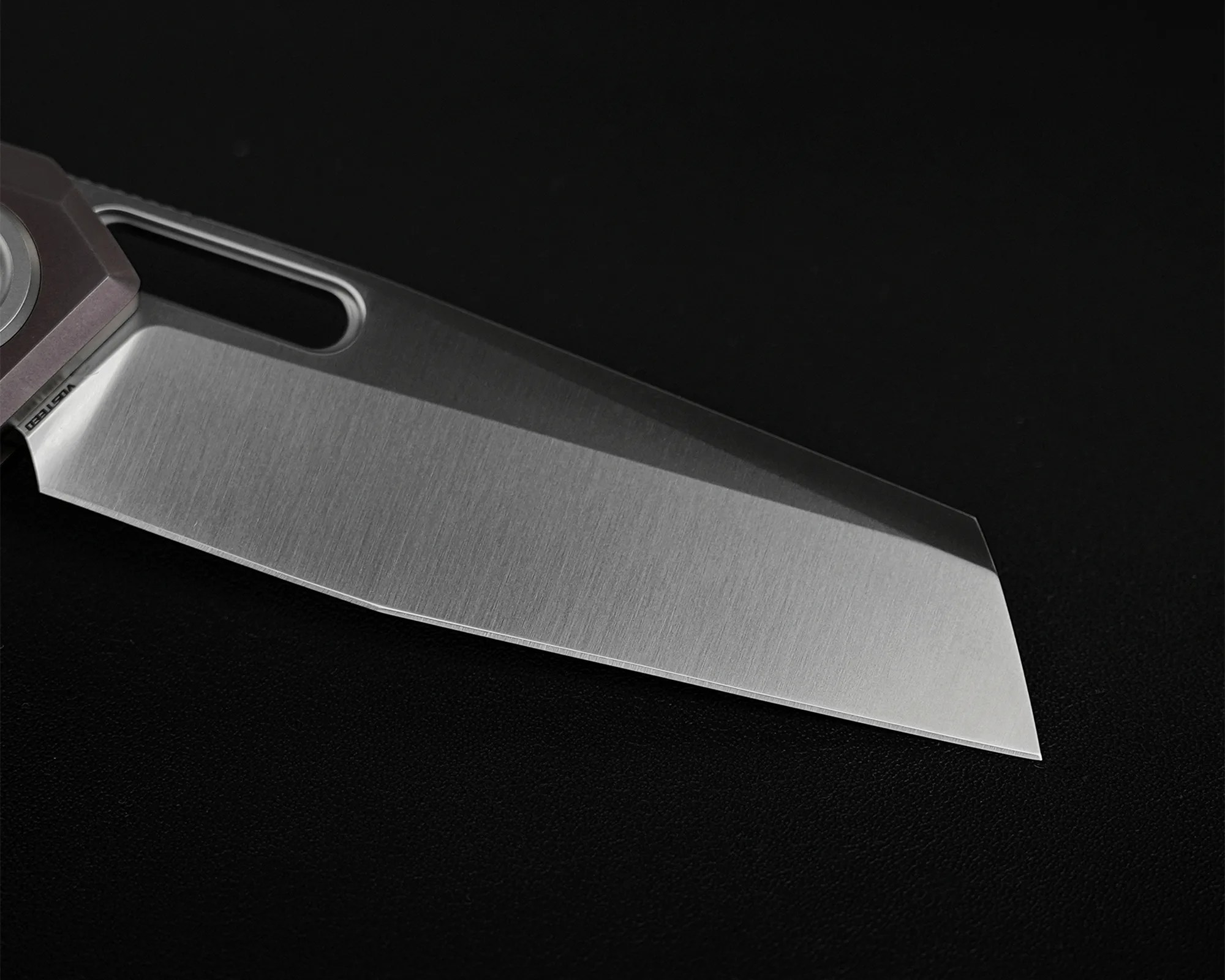 Close-up of a stainless steel knife blade with a brushed finish on a black surface.