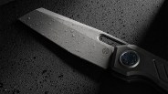 Close-up of a wet, gray folding knife blade with a circular pivot and textured handle on a dark surface.