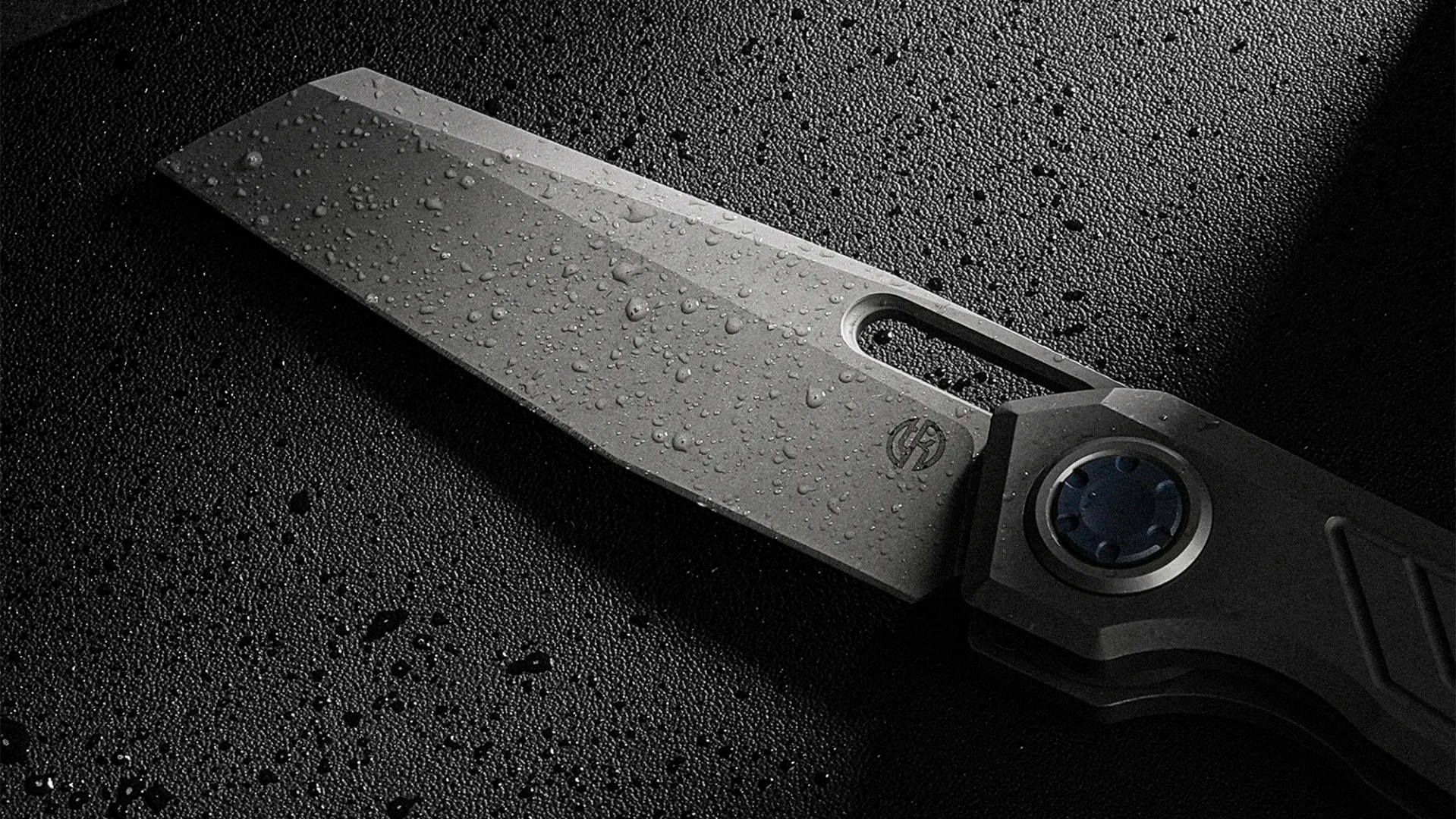 Close-up of a wet, gray folding knife blade with a circular pivot and textured handle on a dark surface.
