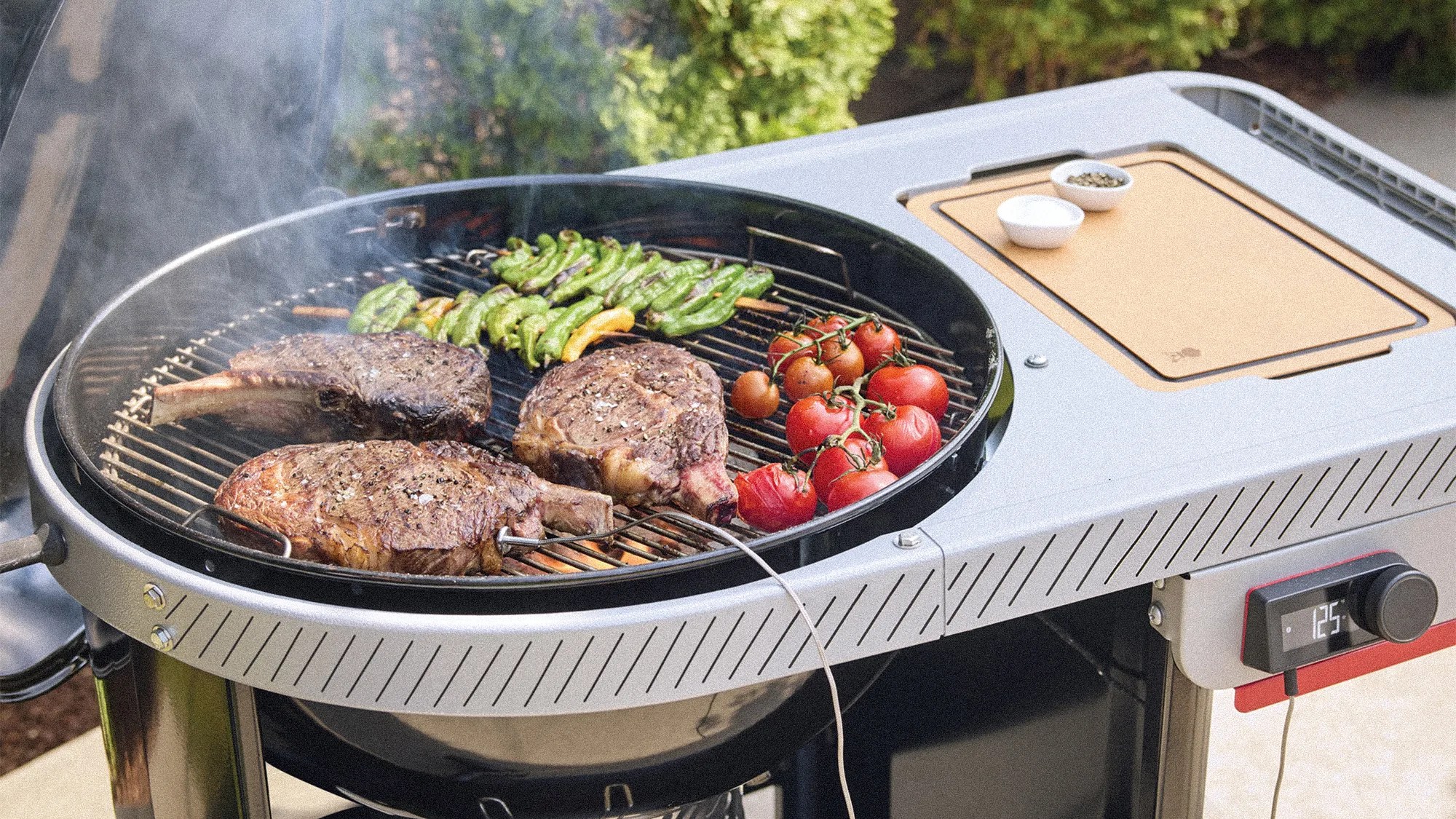 Round charcoal grill with three steaks, green peppers, and vine tomatoes cooking, digital temperature display showing 125&deg;F.