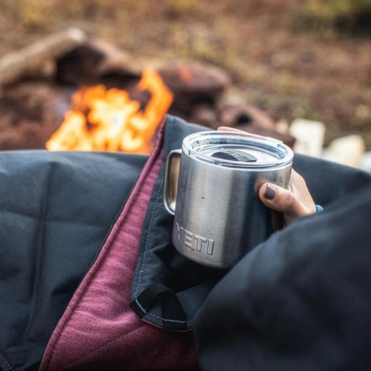 Person holding a stainless steel YETI mug with a clear lid near a campfire, wrapped in a black and red blanket.
