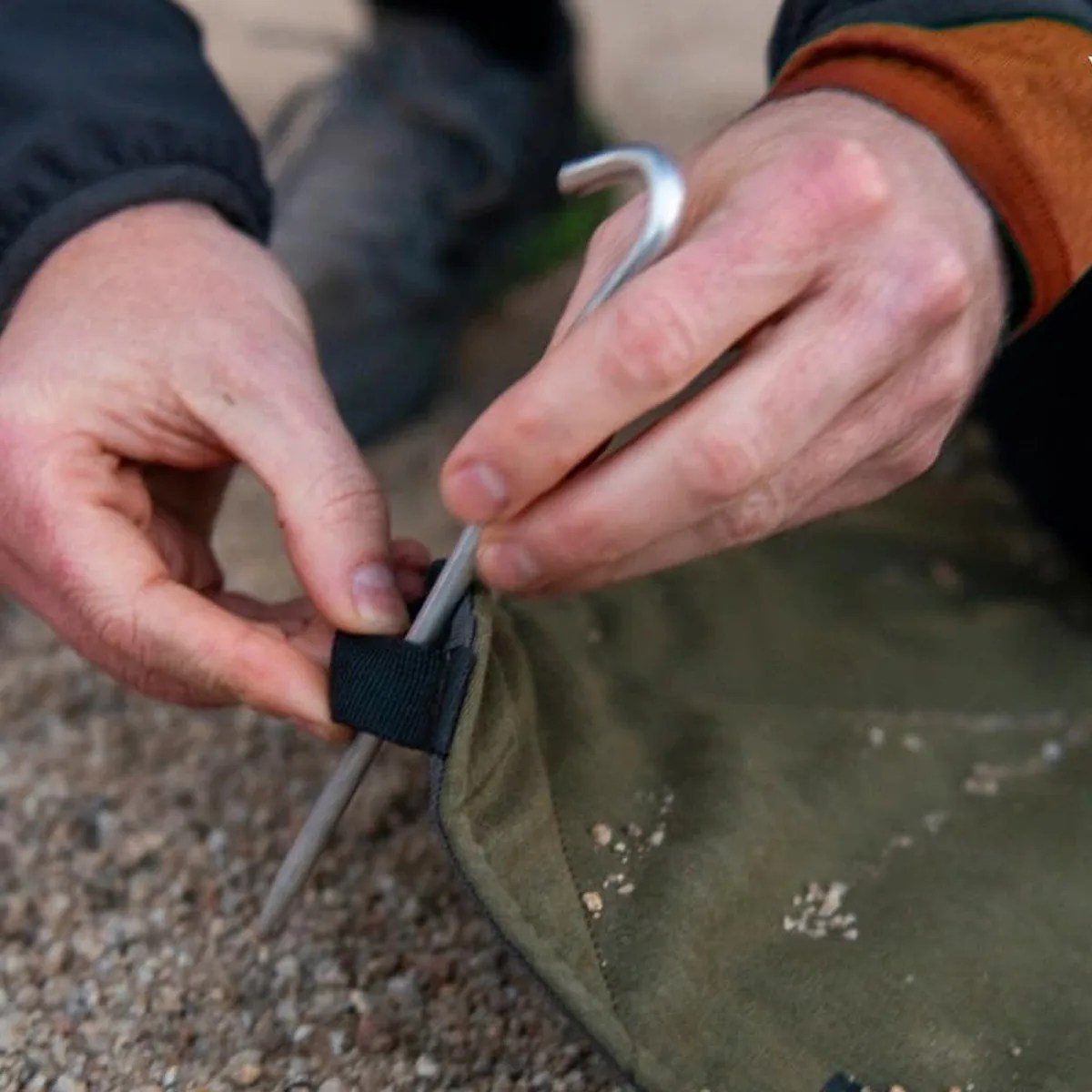 Hands using a metal tent peg to secure a green fabric corner with a black loop on gravel ground.