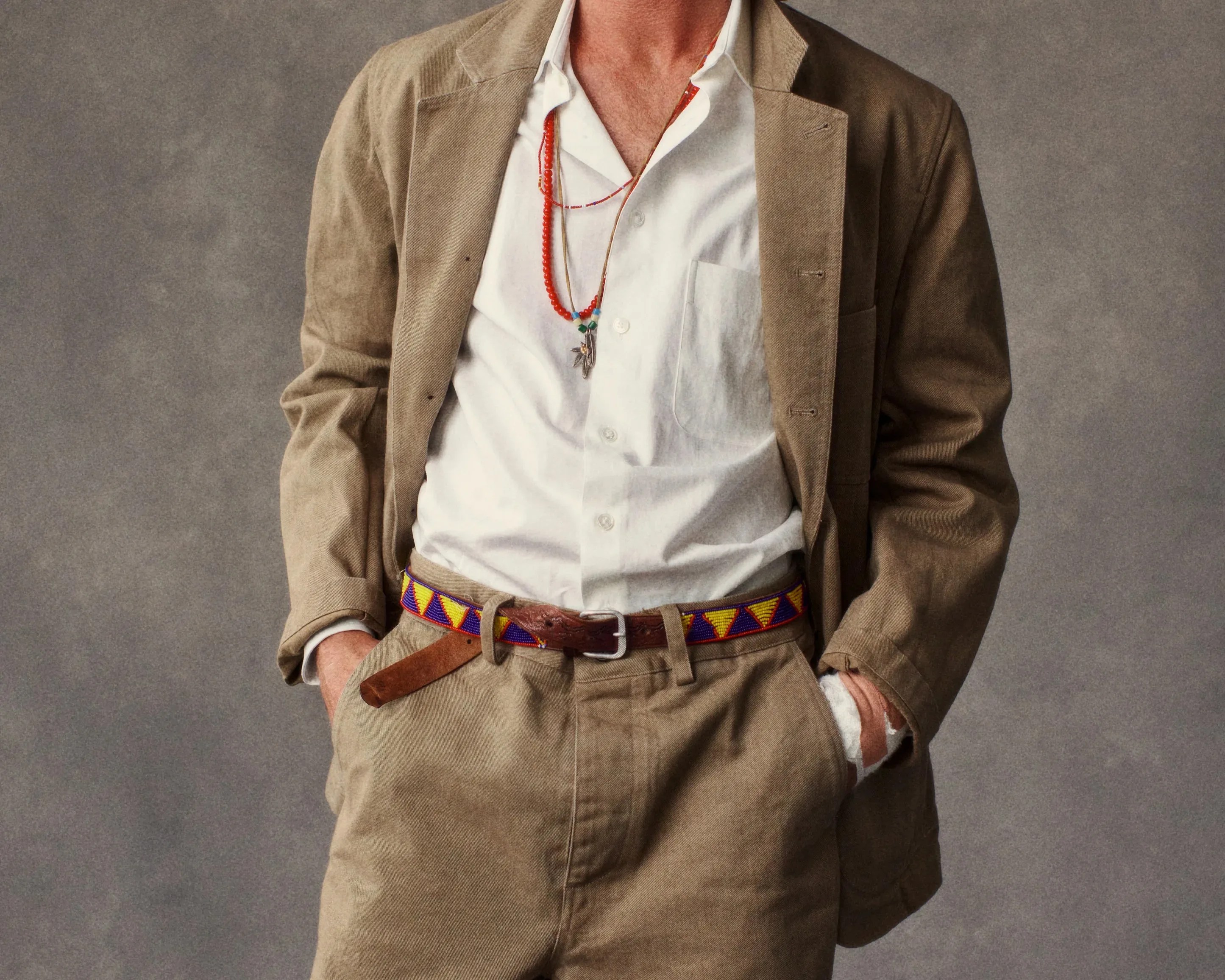 Tan suit with a white button-up shirt, colorful beaded belt, and layered red and silver necklaces.