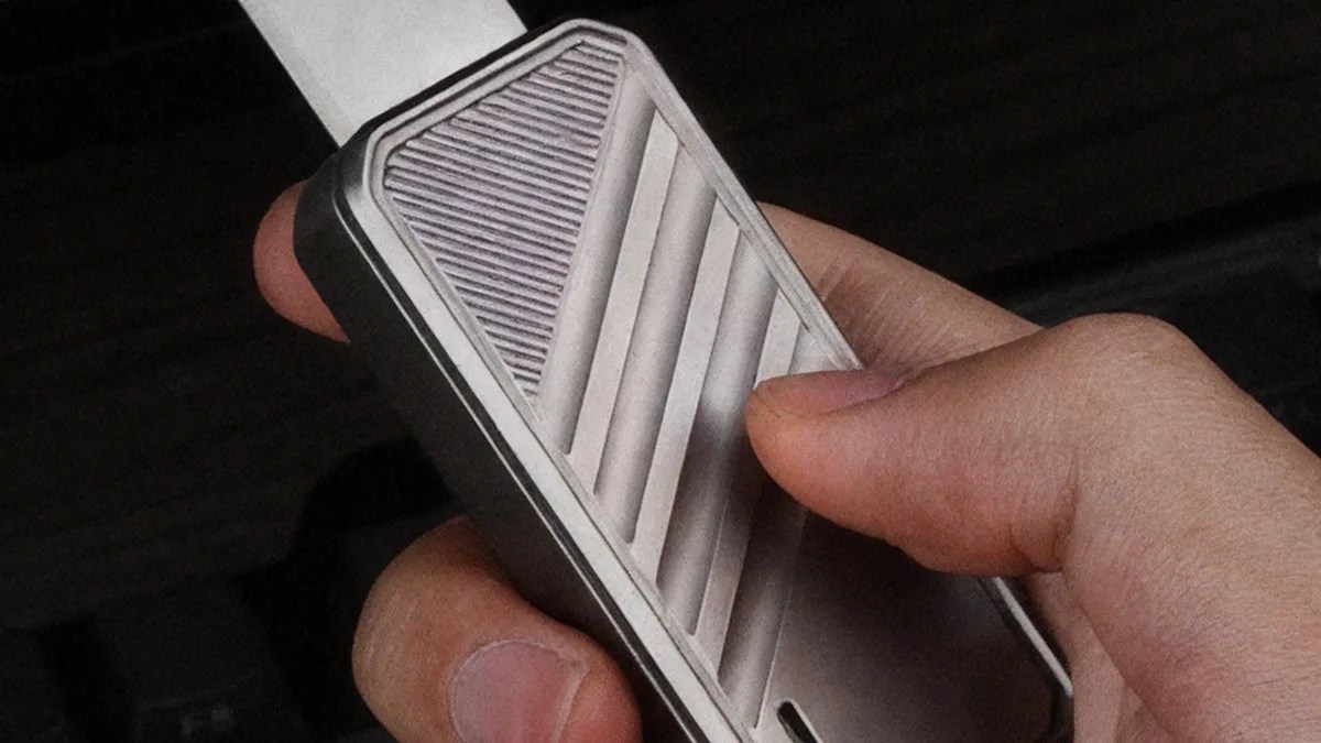 Hand holding a silver rectangular device with diagonal ridges and a white strap attached.