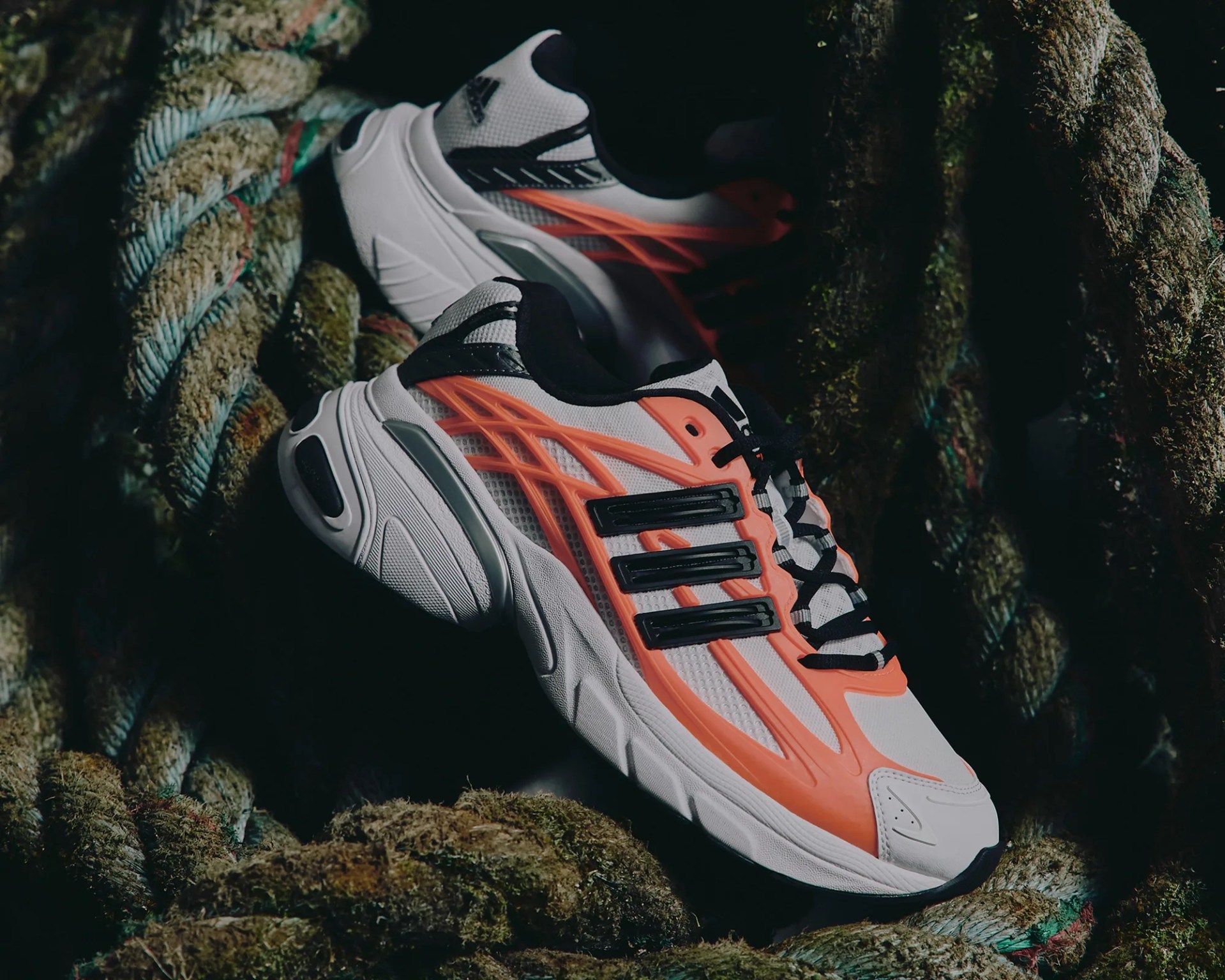 White and orange Adidas sneakers with black stripes and laces resting on thick, moss-covered ropes.
