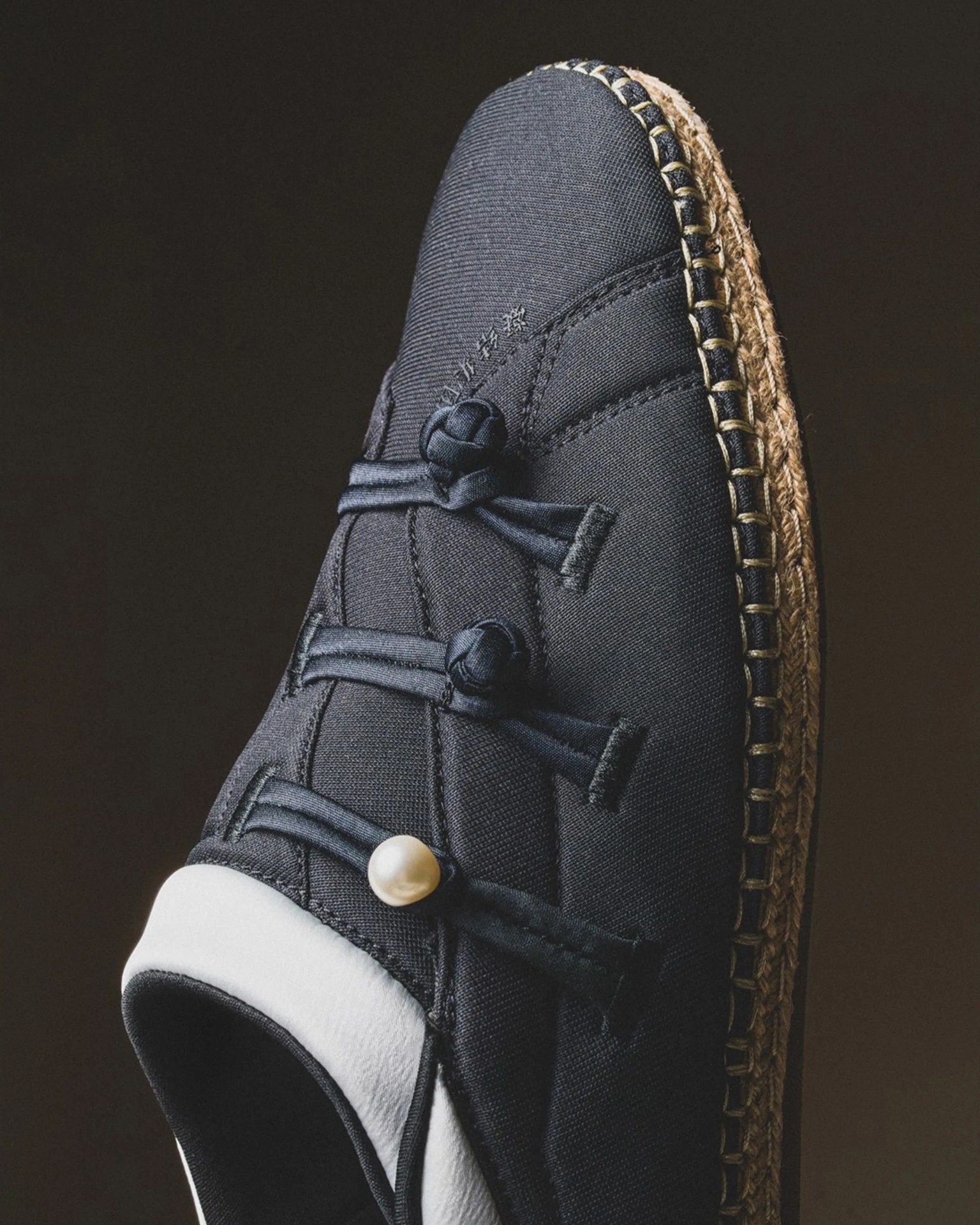 Black fabric shoe with decorative knotted straps and a pearl detail on a woven sole.
