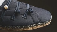 Close-up of a black fabric shoe with knotted laces, a pearl detail, and woven jute sole stitching.