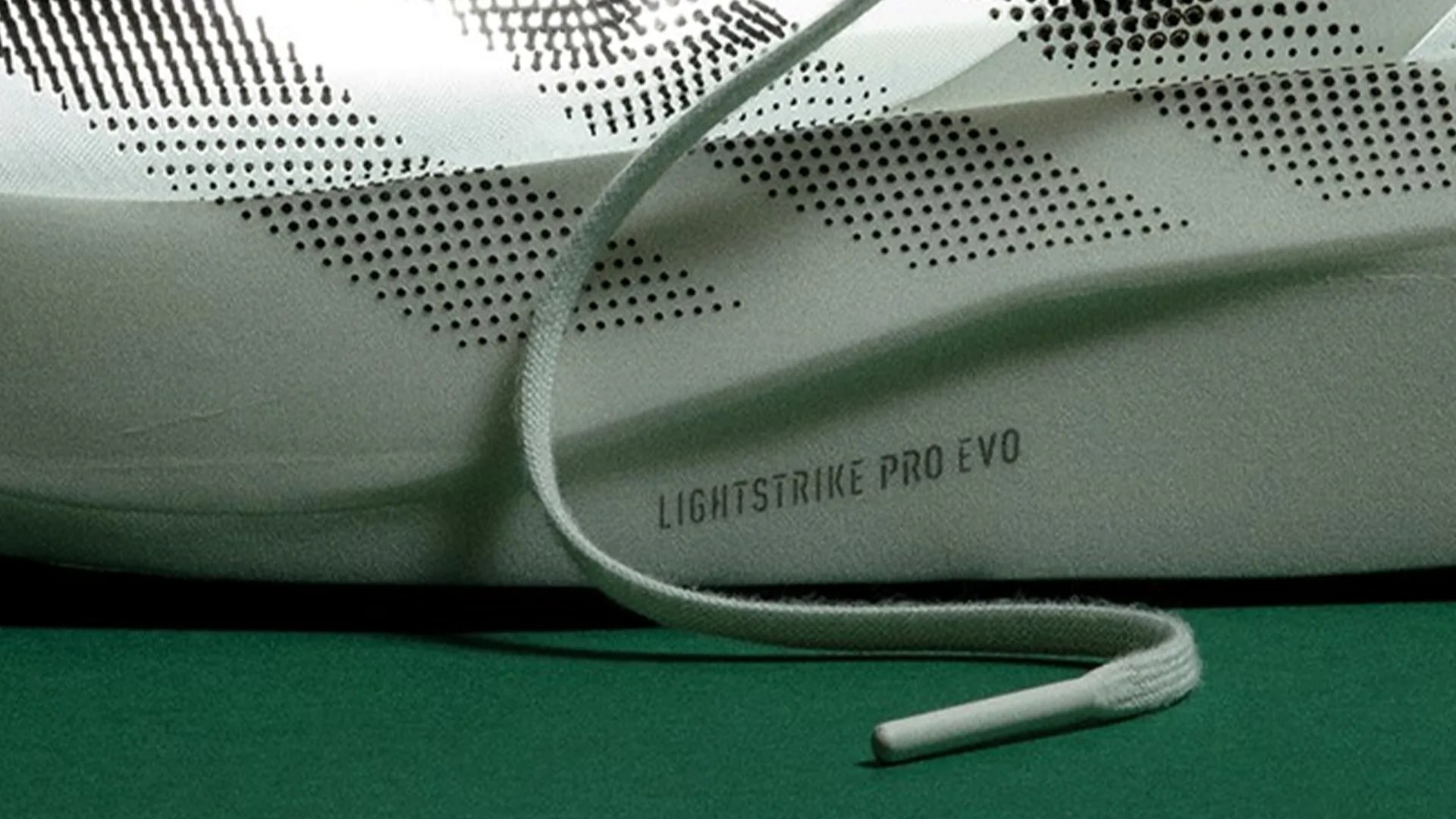 Close-up of a white sneaker with perforated triangular patterns and "LIGHTSTRIKE PRO EVO" text, with a white shoelace on a green surface.