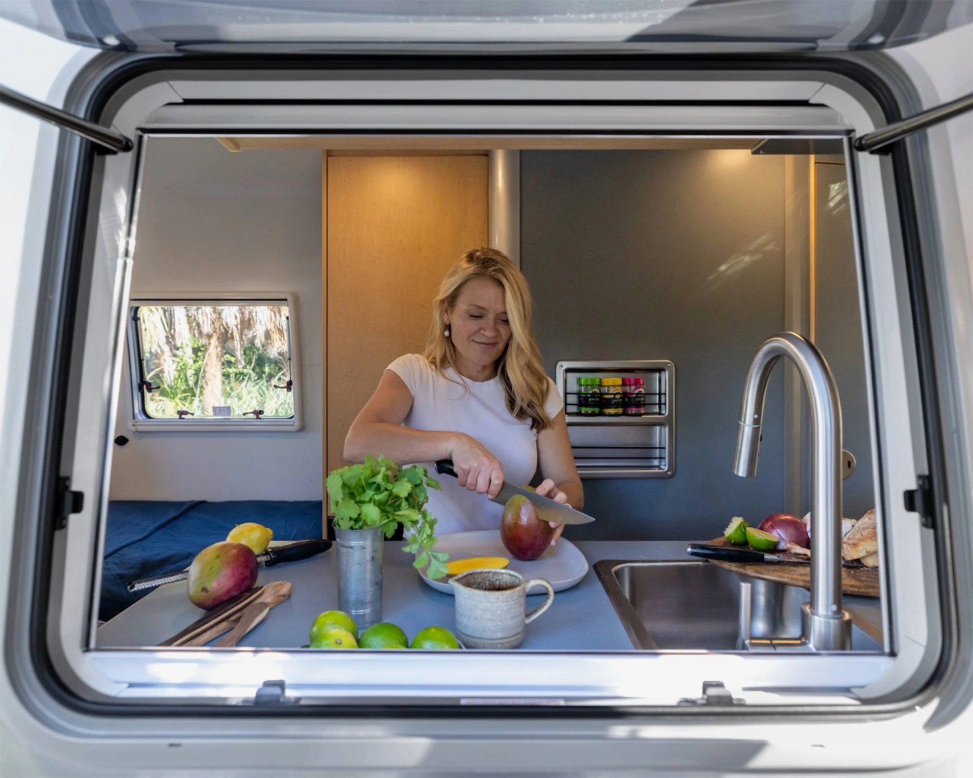 Woman slicing a mango in a compact camper kitchen with a stainless steel faucet and fresh produce on the counter.