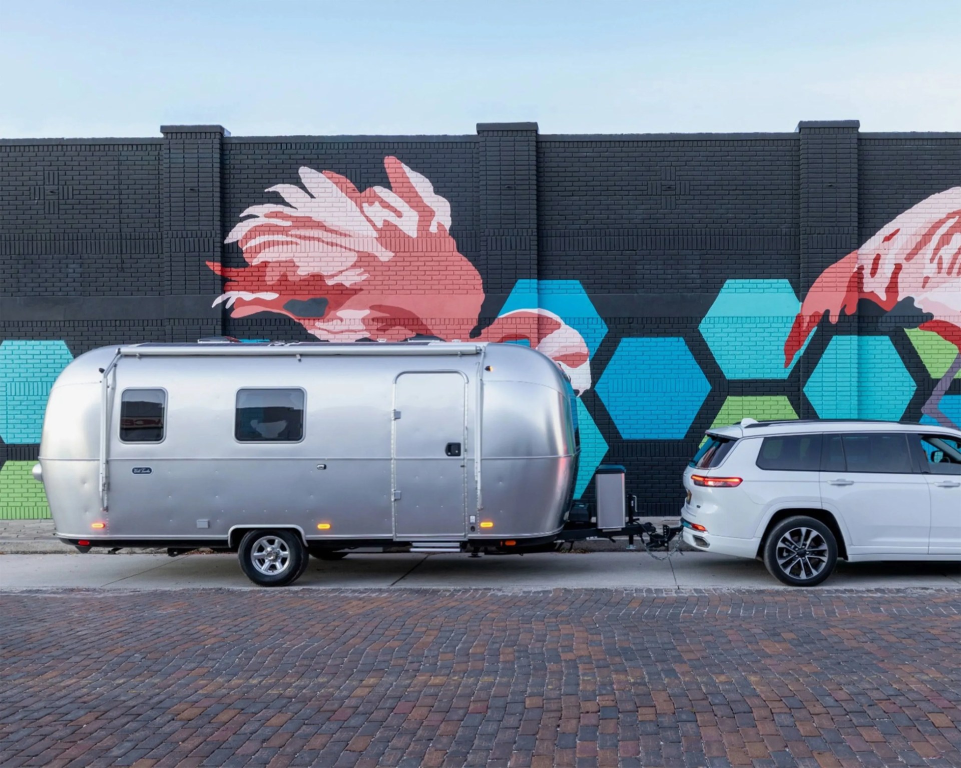 Silver Airstream trailer hitched to a white SUV parked in front of a black brick wall with colorful fish mural.