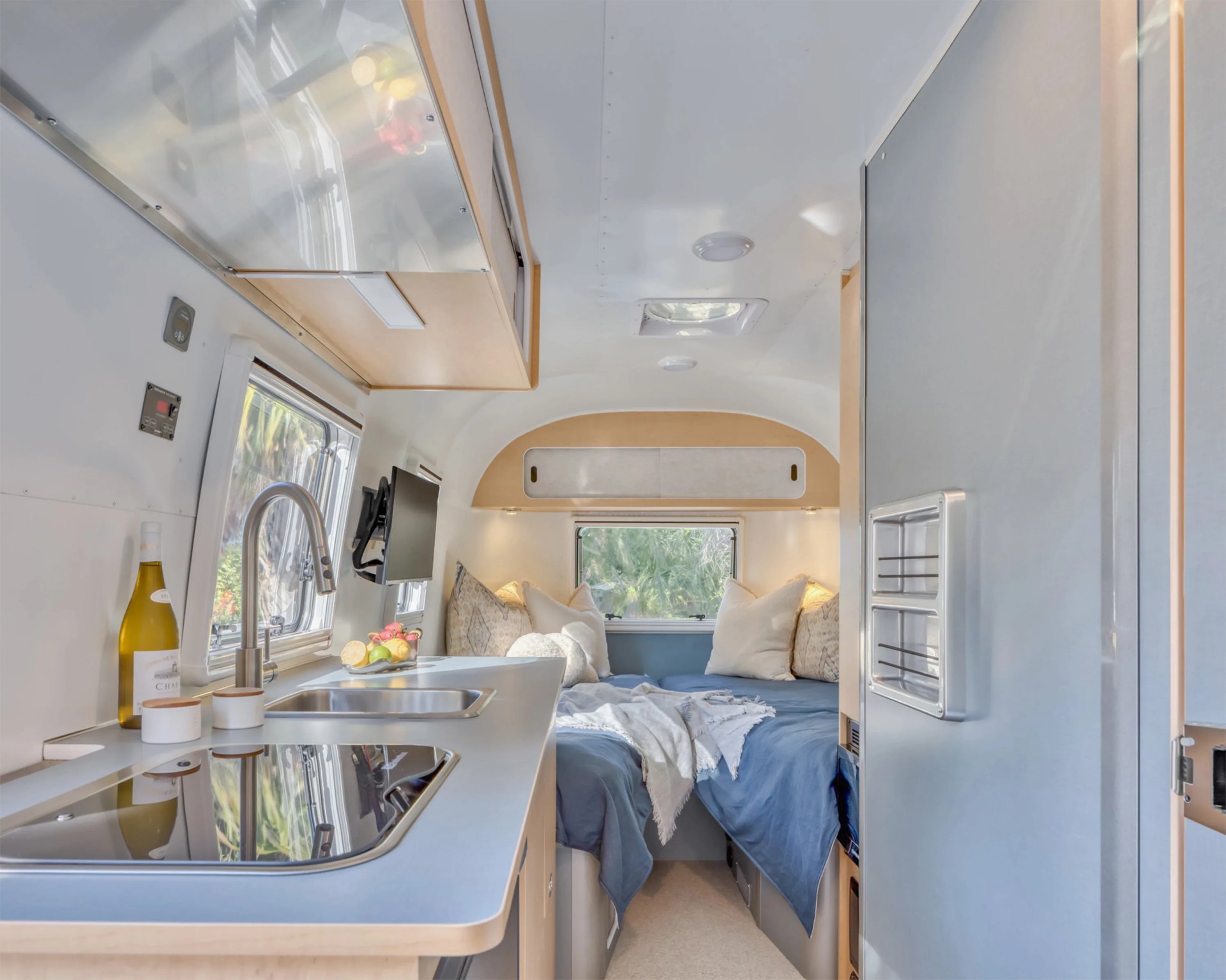 Compact camper interior with a kitchenette featuring a sink, stovetop, and a bed with blue bedding and pillows at the rear.