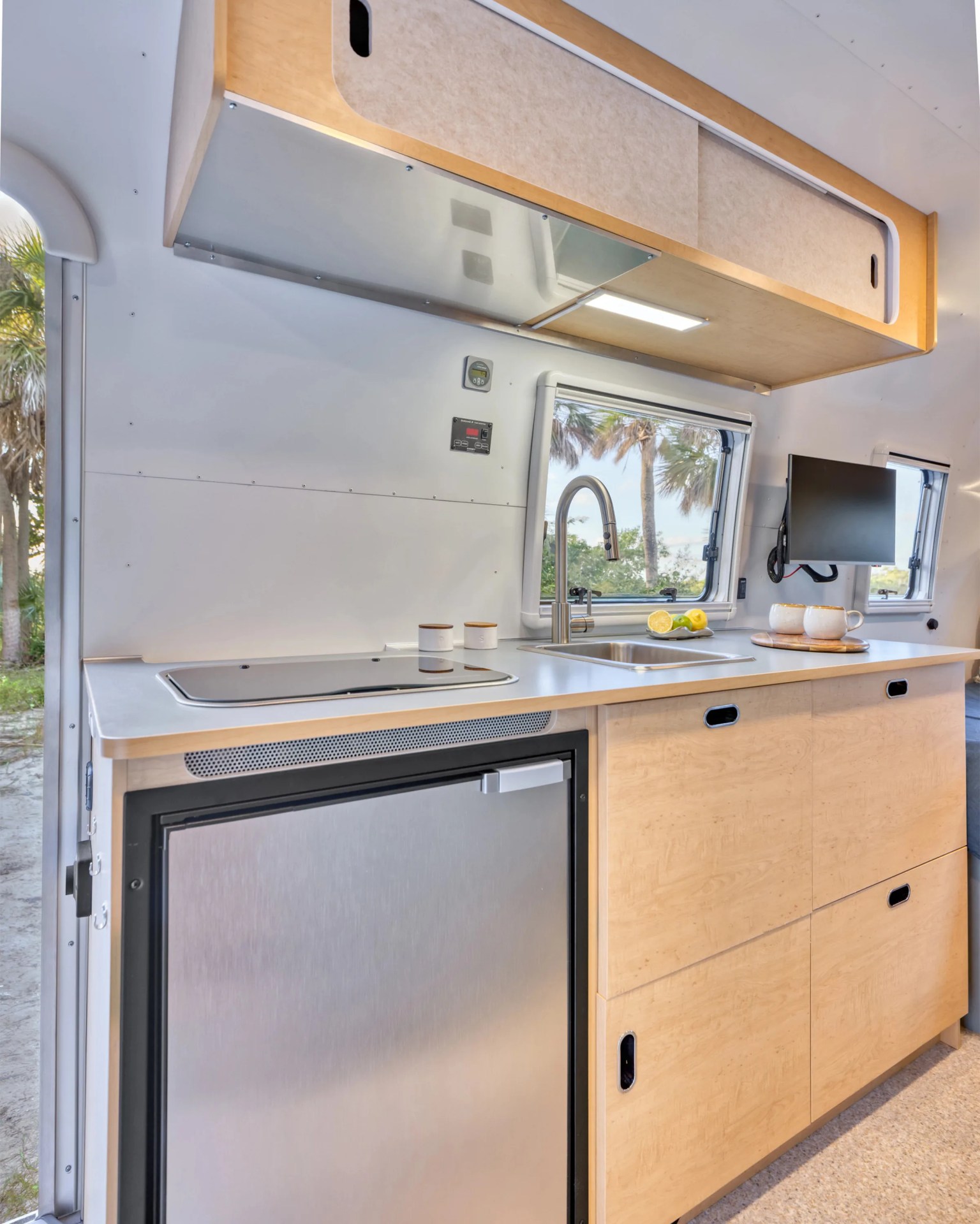 Compact RV kitchen with light wood cabinets, stainless steel mini fridge, sink, and window showing palm trees.