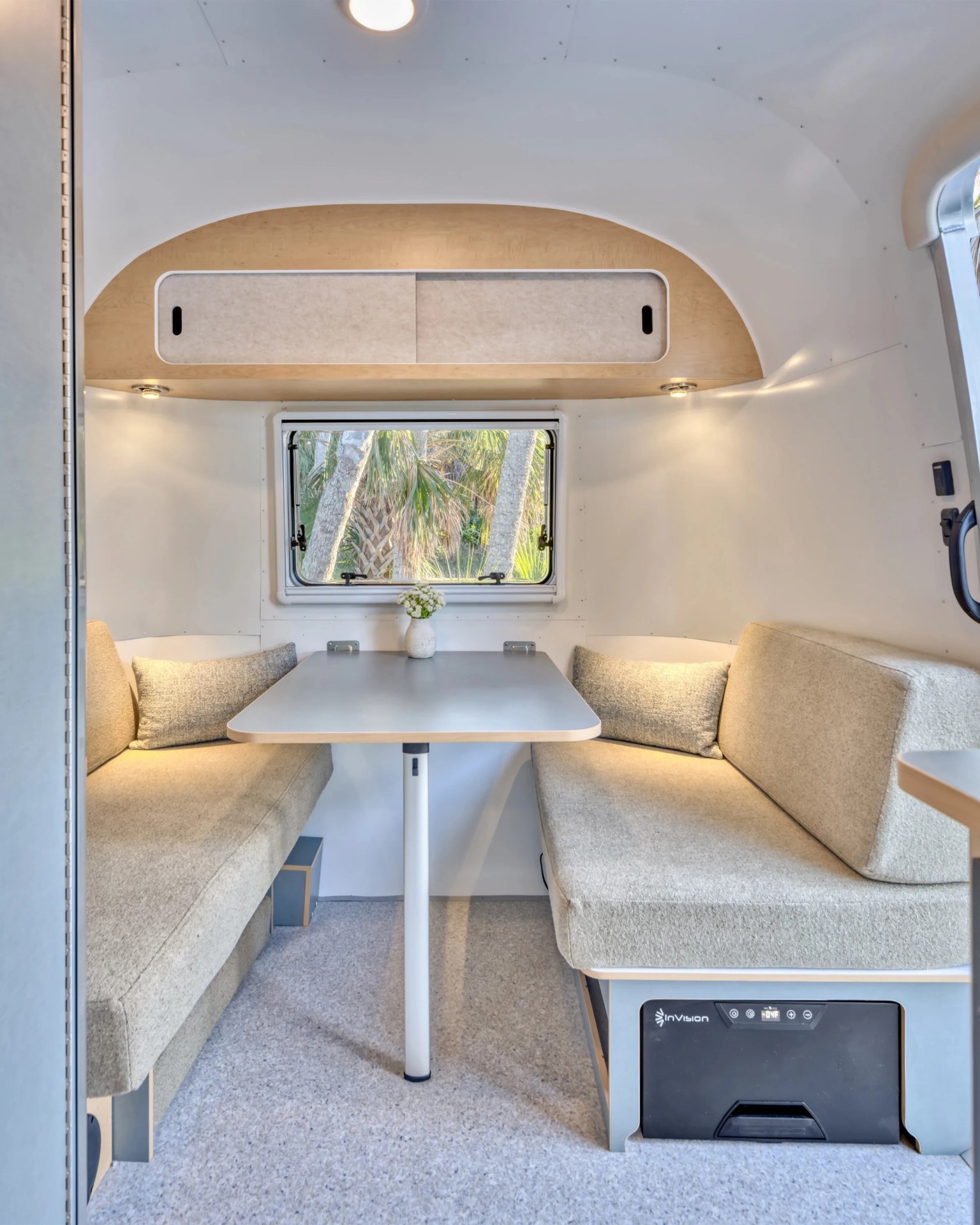 Compact camper dining area with beige cushioned benches, a white table, overhead storage, and a window showing palm trees.