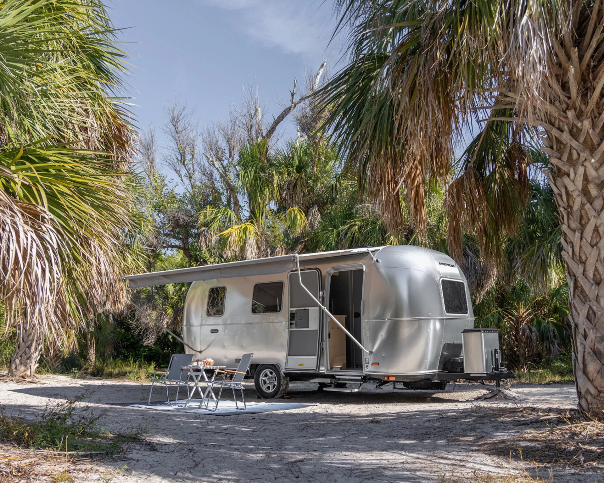 Silver Airstream trailer with extended awning and outdoor table and chairs set on sandy ground surrounded by palm trees.