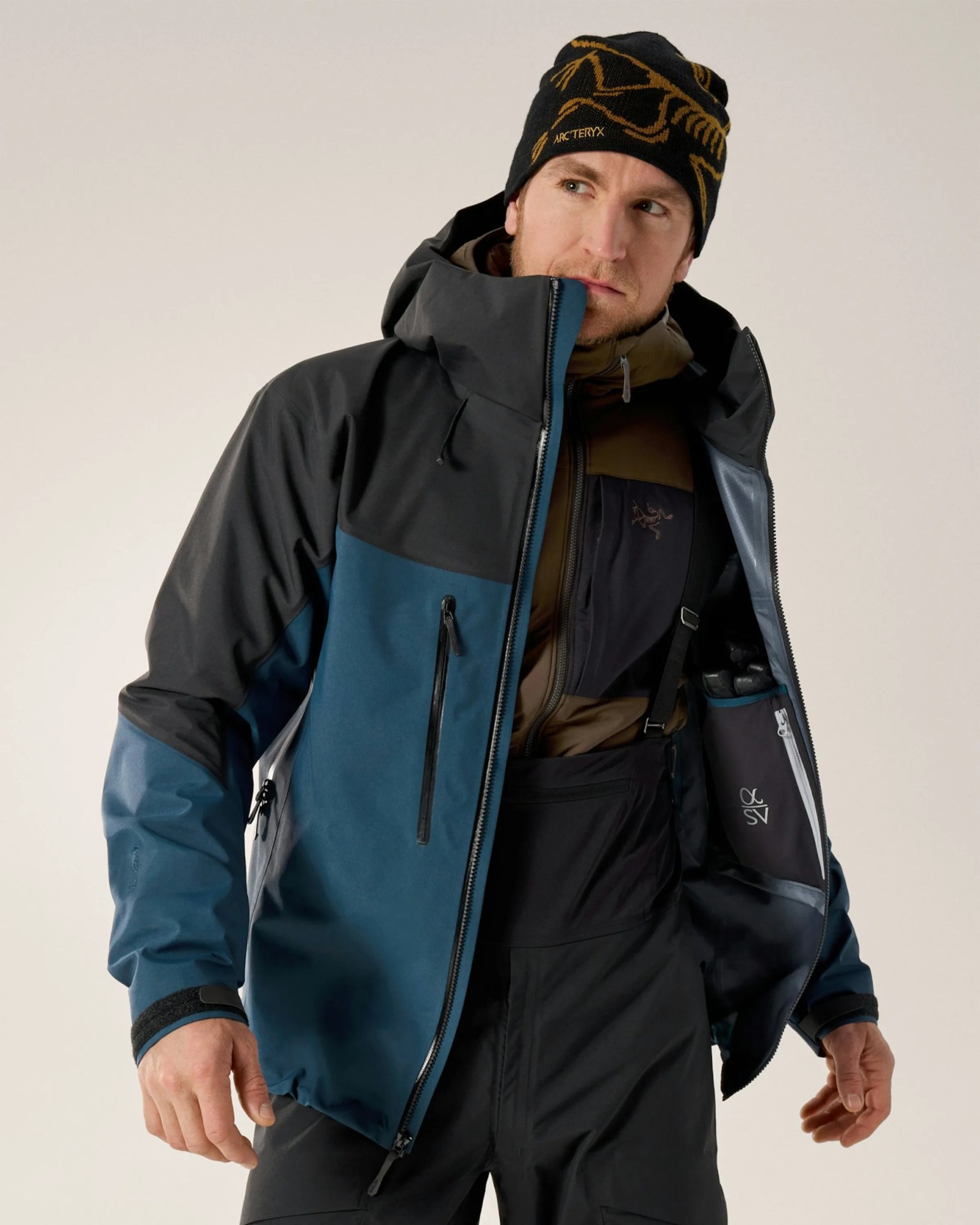 Man wearing a blue and black zip-up jacket with a hood and a black and gold Arc'teryx beanie.