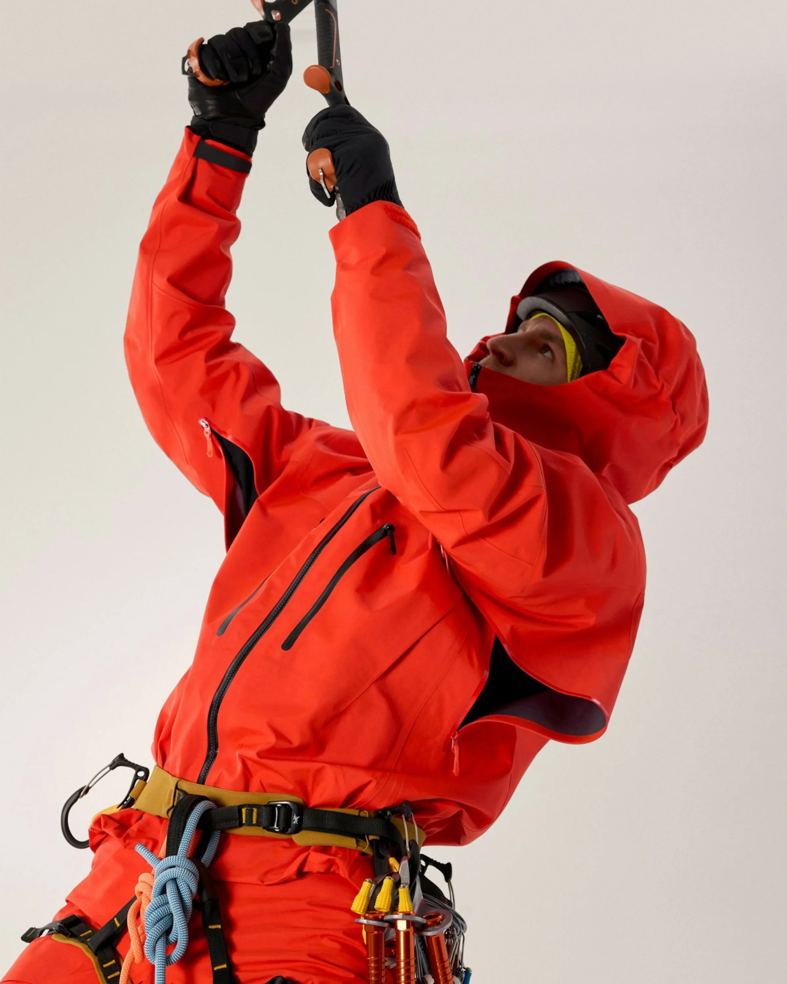 Person wearing a bright red waterproof jacket with hood and black gloves, holding climbing gear attached to a harness.