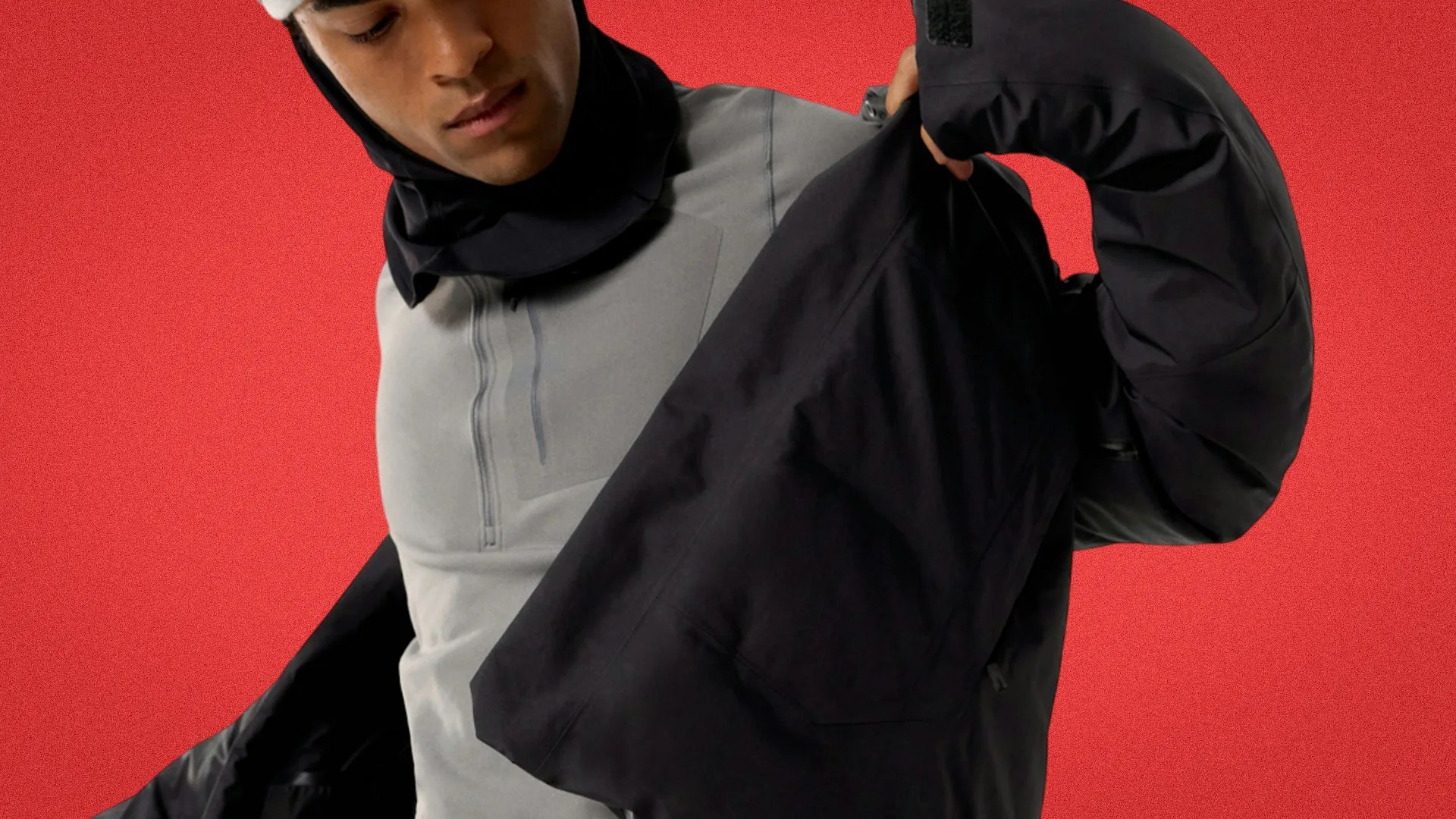 Person wearing a light gray zip-up top and putting on a black jacket against a red background.