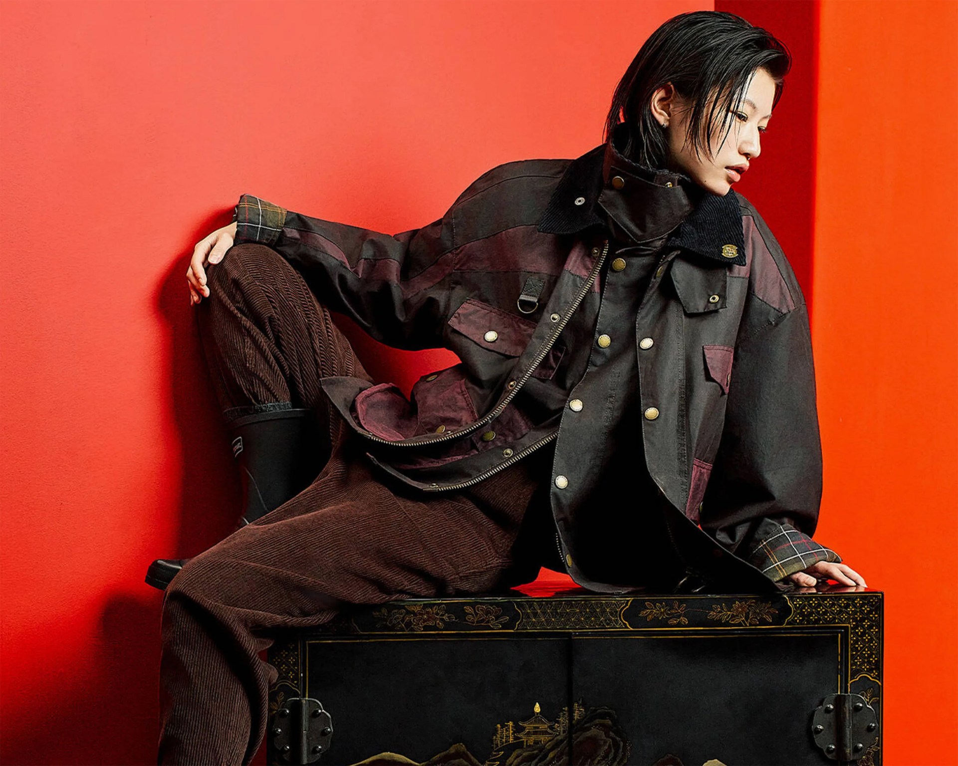 Person wearing a dark brown and black jacket with brass buttons and brown corduroy pants sitting against a red wall.