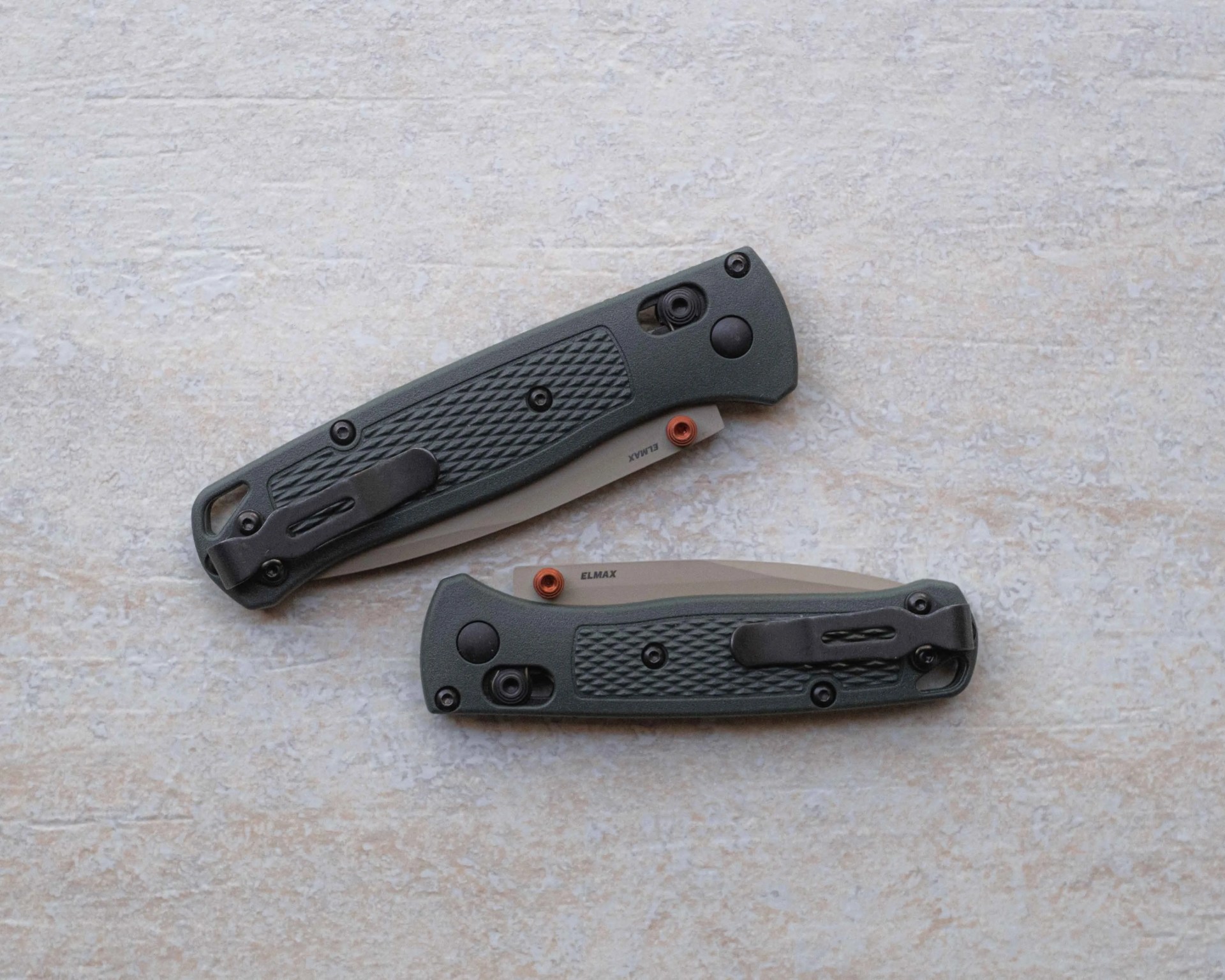 Two closed folding knives with dark textured handles and partially visible blades labeled "ELMAX" on a light stone surface.