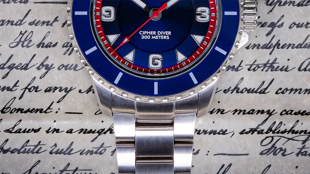 Close-up of a silver metal watch with a blue bezel and dial labeled "Cipher Diver 300 Meters" on a handwritten paper background.