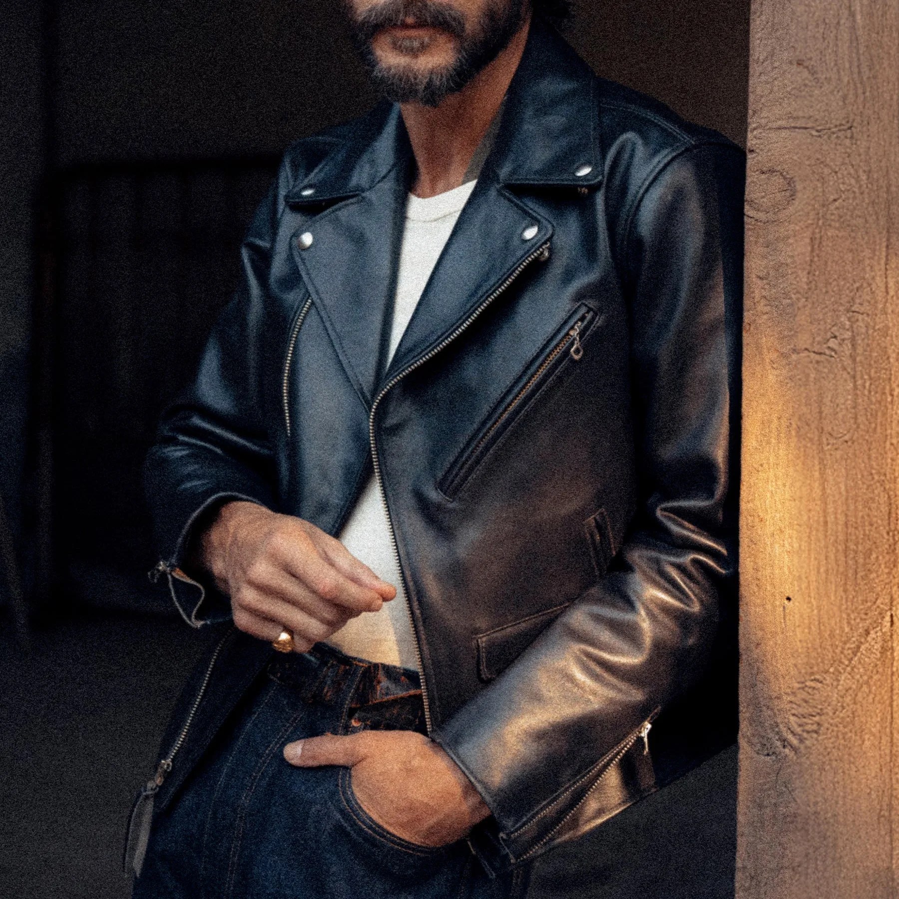 Man wearing a black leather biker jacket over a white shirt with one hand in dark blue jeans pocket.
