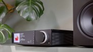 Black audio receiver with a large volume knob and digital display showing a music track, placed on a shelf next to a speaker and green leaves.
