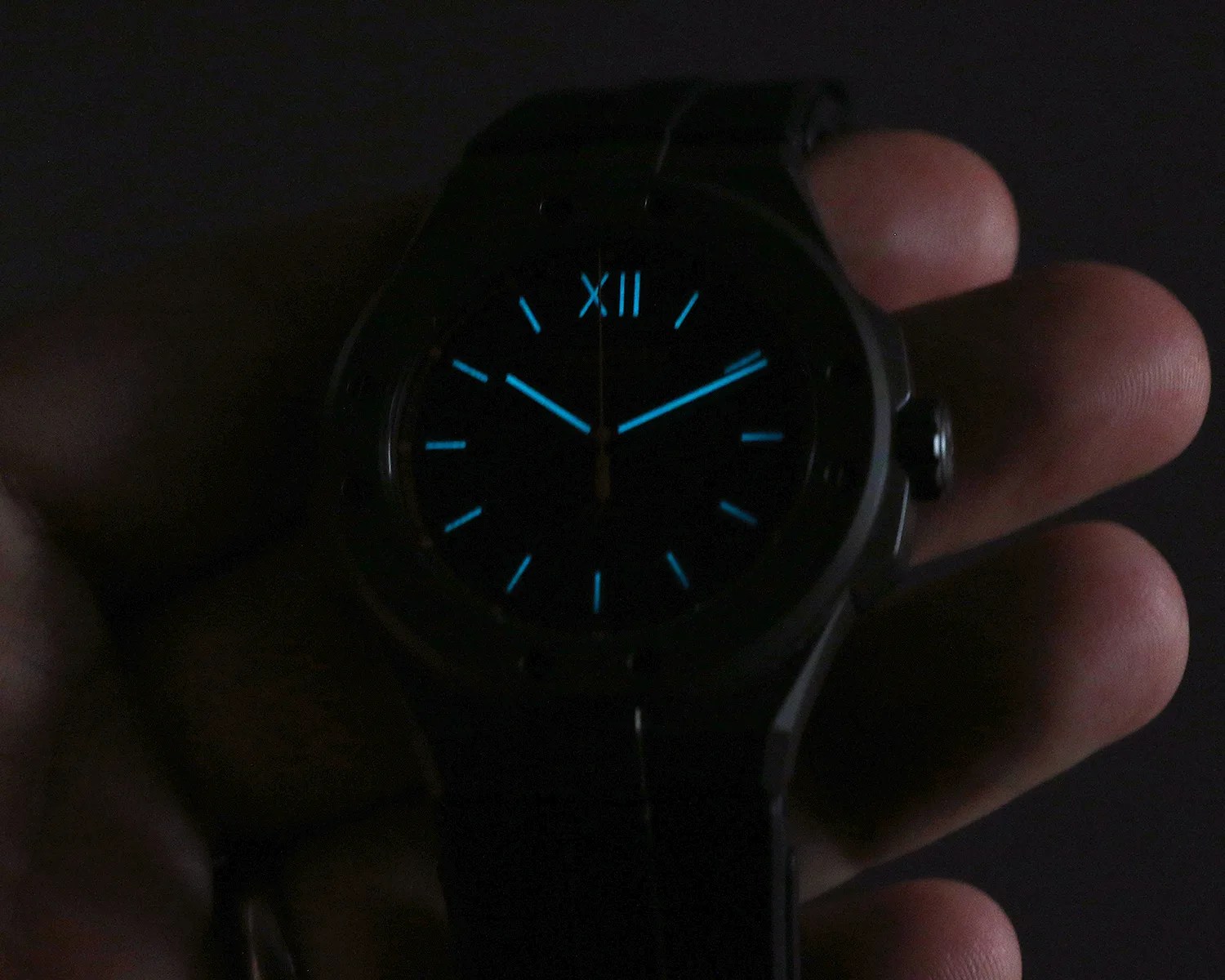 Black wristwatch with glowing blue hour markers and hands held in a hand against a dark background.