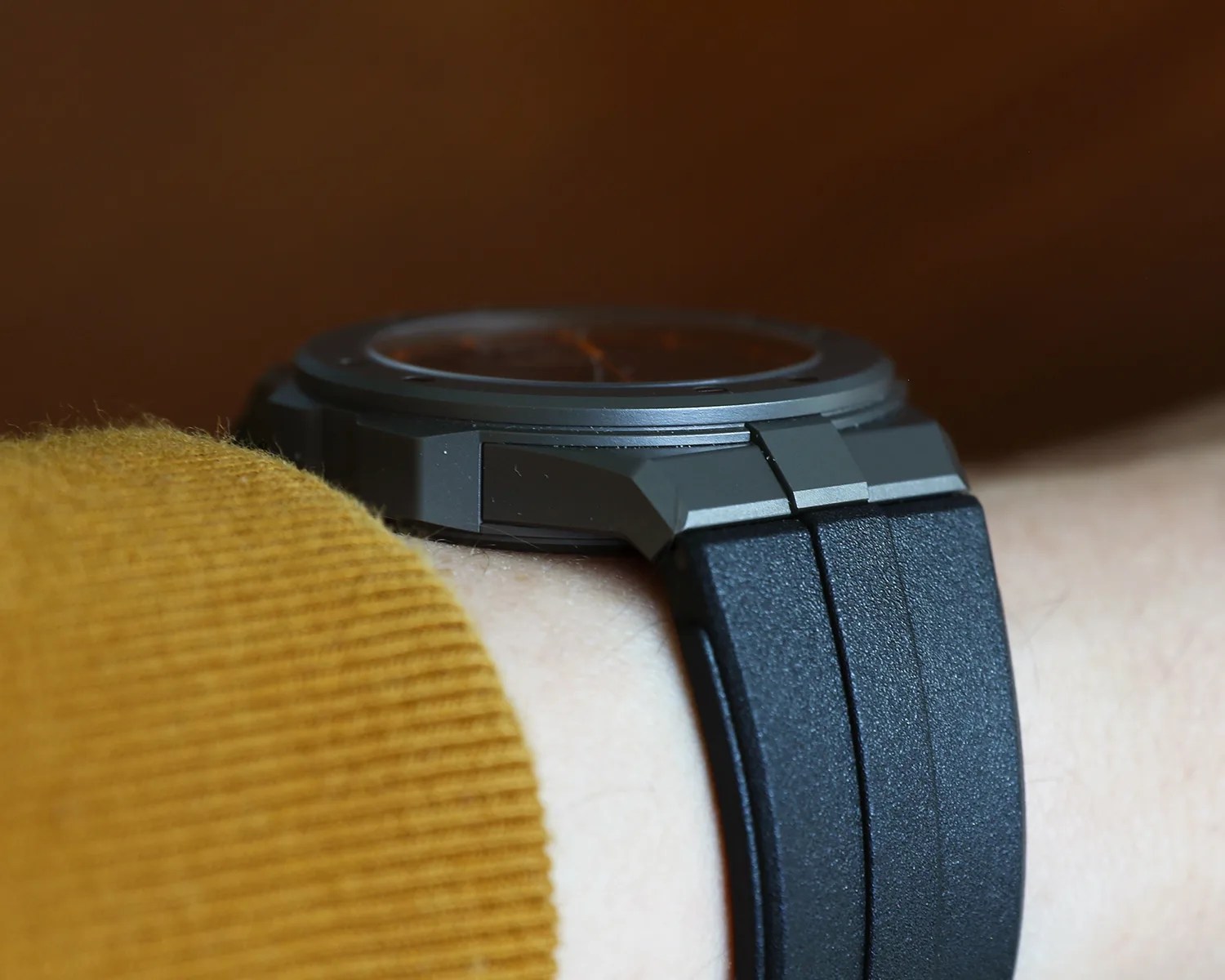 Close-up of a black watch with a textured rubber strap worn on a wrist with a mustard-colored sleeve.