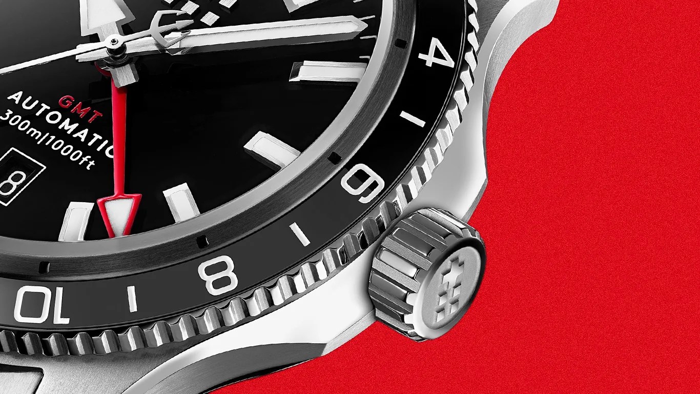 Close-up of a black GMT automatic watch face with white markers, a red GMT hand, and a silver crown on a red background.