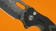 Close-up of a folding knife with a patterned Damascus steel blade and a black textured handle on an orange background.