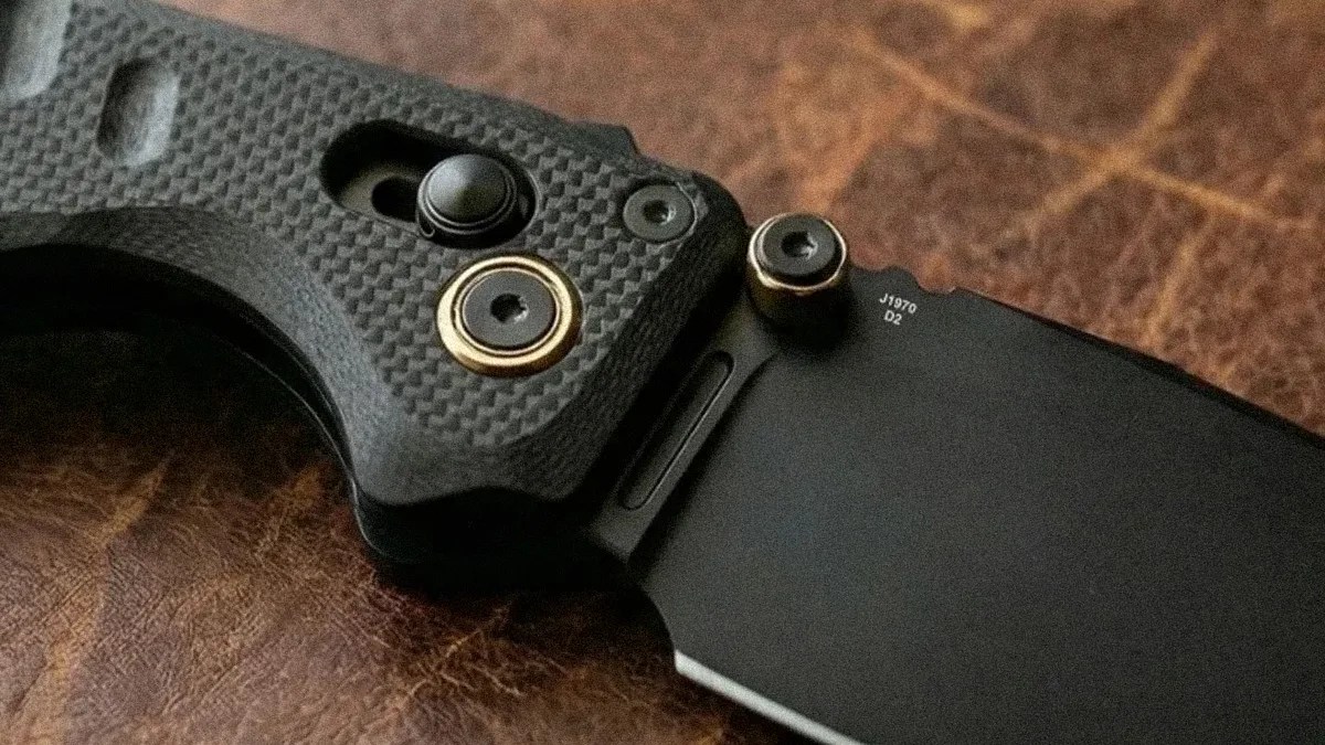 Close-up of a black folding knife with textured handle and gold-accented pivot screws on a brown leather surface.